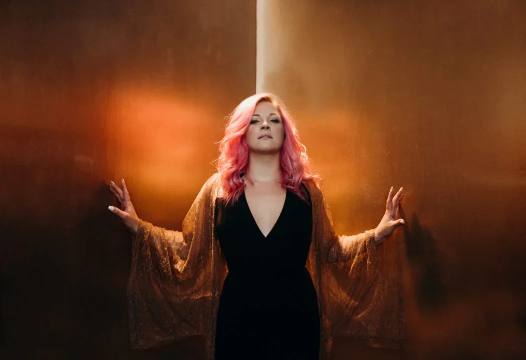 A woman with pink hair leaning against a warm-toned wall, wearing a black dress with a deep neckline and a brown shawl.