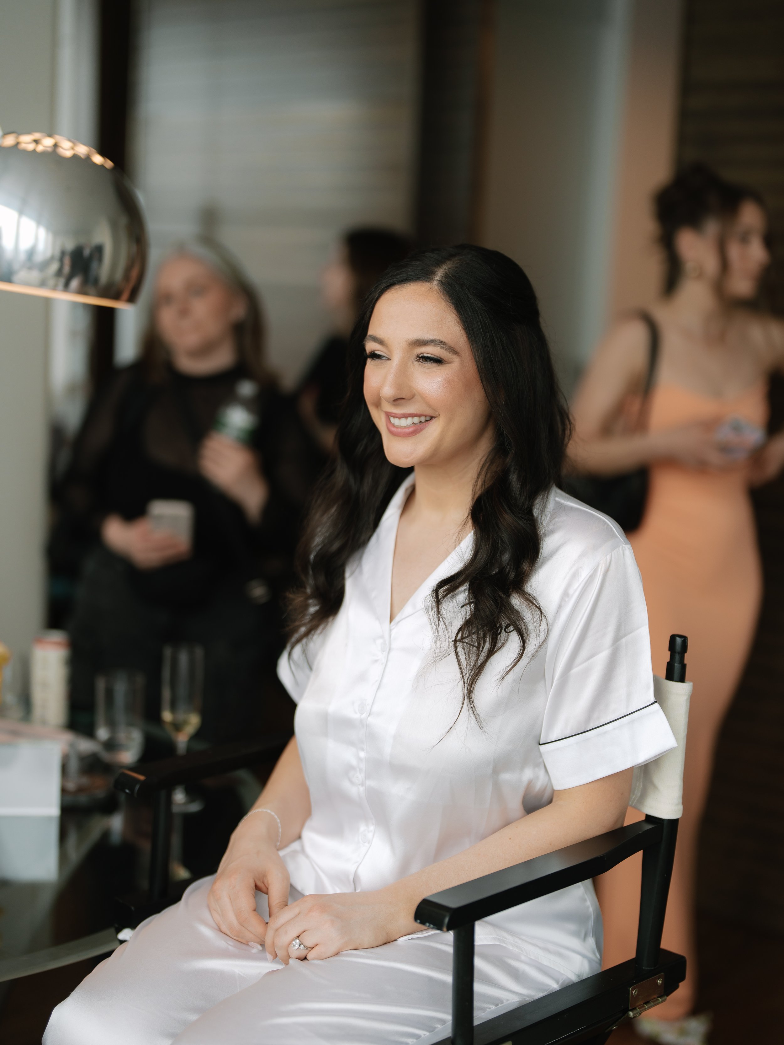 A woman smiling in satin pajamas sitting in a makeup chair at a dressing table with a mirror and makeup items, in a room with other women in the background.