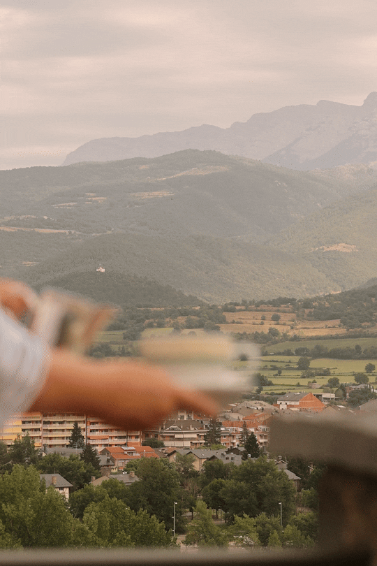 Hospitality lifestyle photography: Blurred foreground of a person pouring tea, overlooking a scenic mountain landscape and town at El Castell de Ciutat. Professional hotel portfolio imagery.
