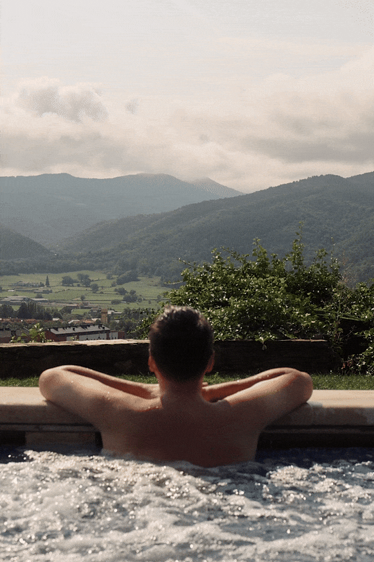 Spa and wellness lifestyle photography: A guest relaxing in an outdoor hot tub with panoramic views of the Catalan Pyrenees valley. Professional hospitality portfolio imagery at El Castell de Ciutat.