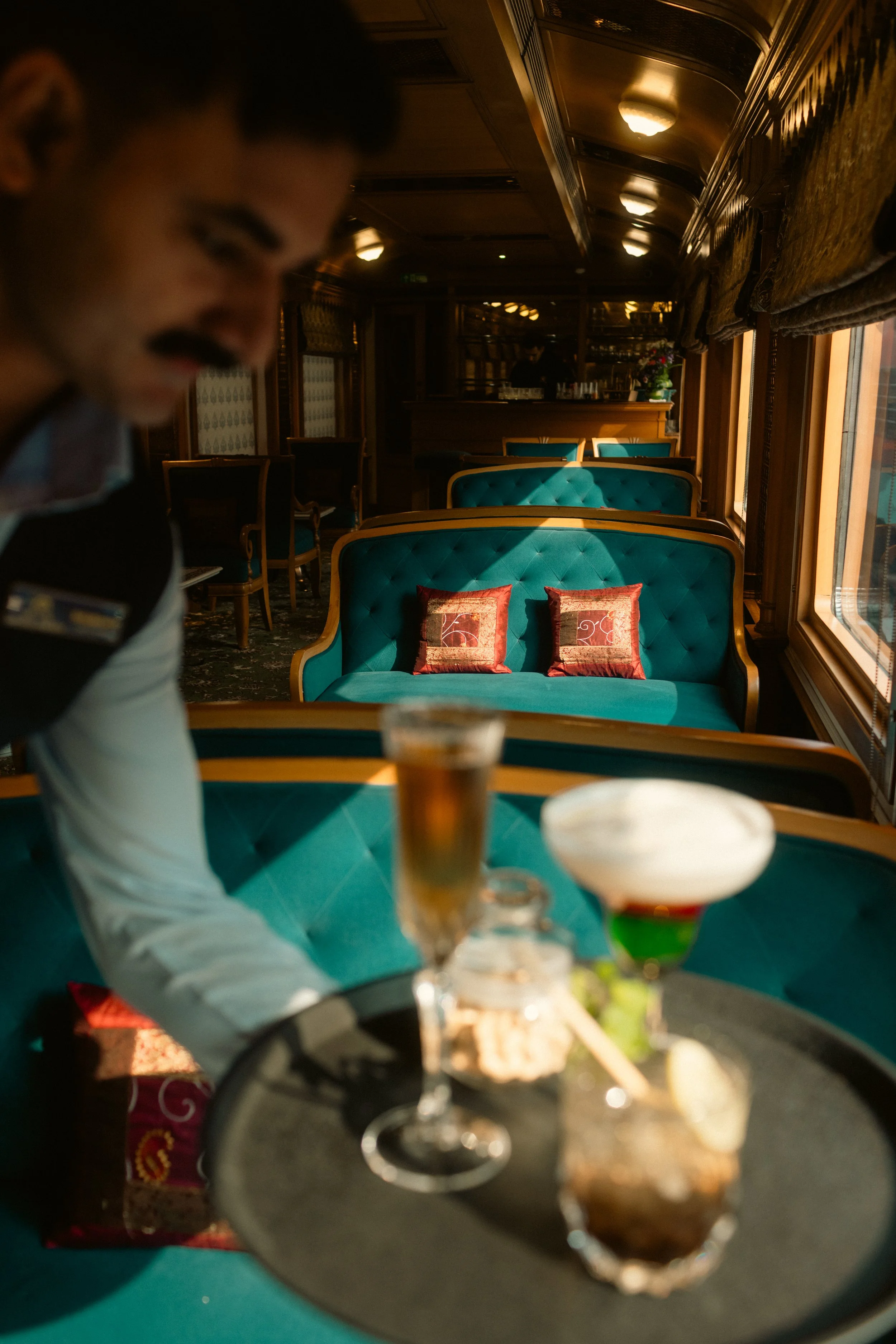 Hospitality lifestyle photography: A server holding a tray with cocktails in a vintage-style lounge with teal seating. Professional bar and service portfolio imagery at Deccan Odyssey.