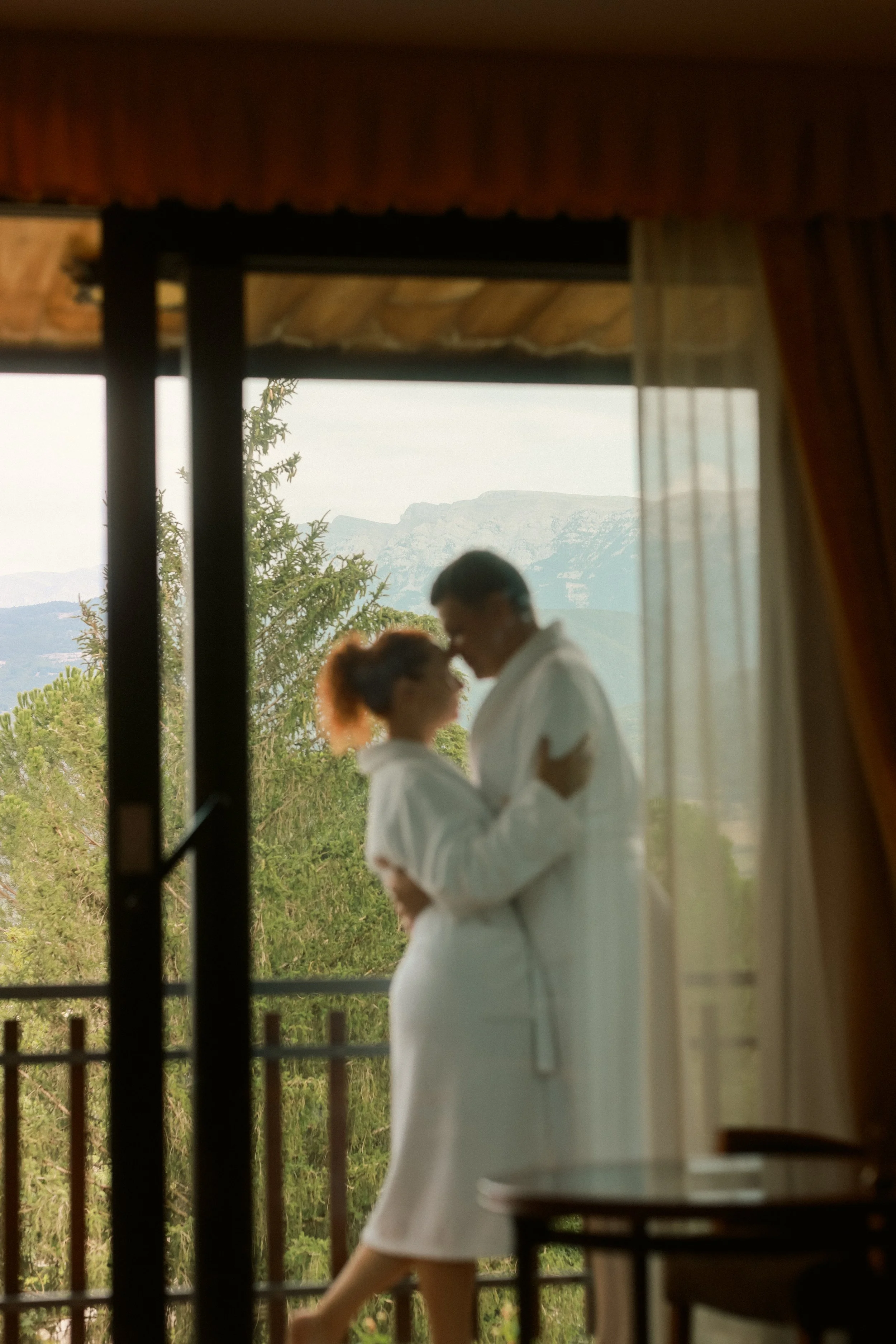 Luxury hotel lifestyle photography: A couple in white robes embracing on a balcony with a mountain backdrop, framed by curtains at El Castell de Ciutat. Professional hospitality portfolio imagery.
