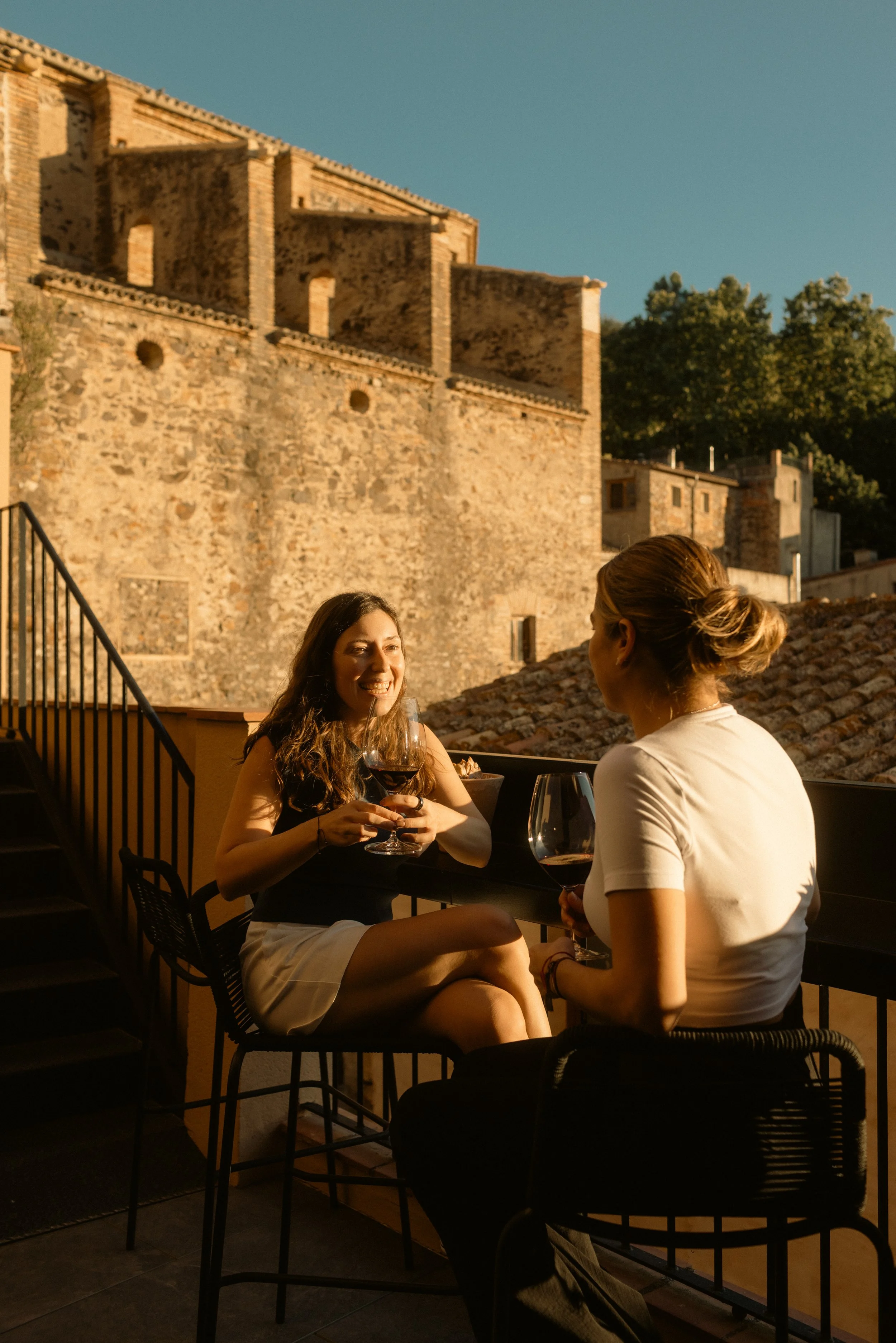 ORA Priorat hotel lifestyle photography, two guests enjoying wine on a balcony at sunset with historic stone buildings and surrounding greenery, Catalonia, Spain.
