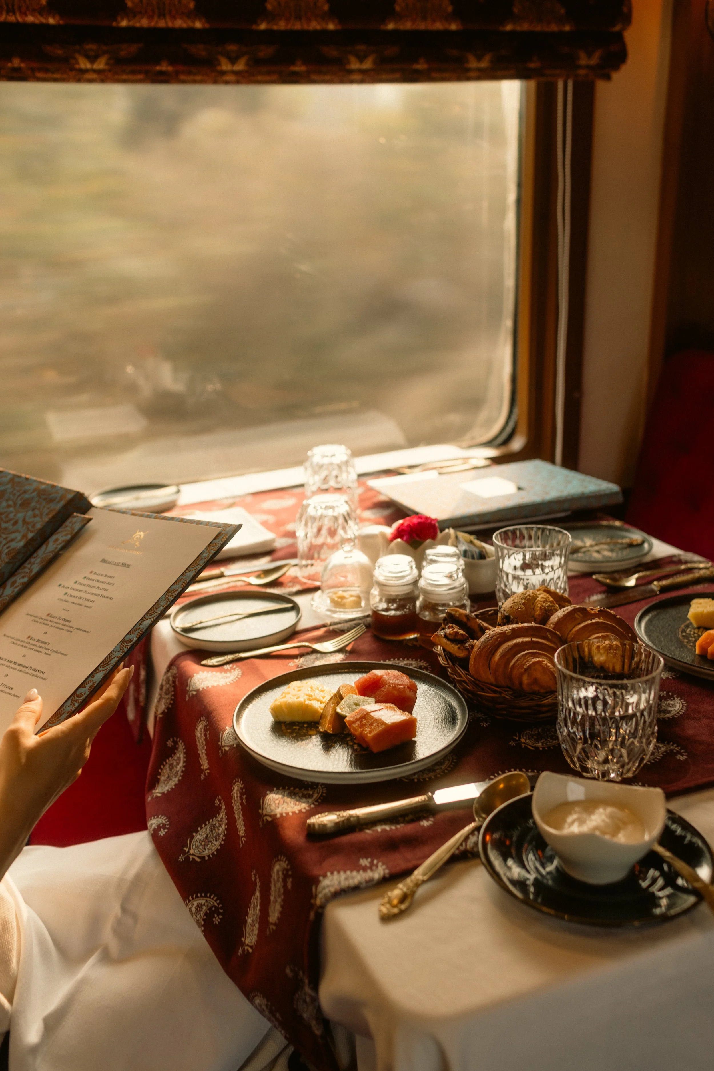 A festive breakfast or brunch table set with various pastries, cheeses, jams, and fruit, with a window in the background showing a blurred view outside and sunlight coming in.