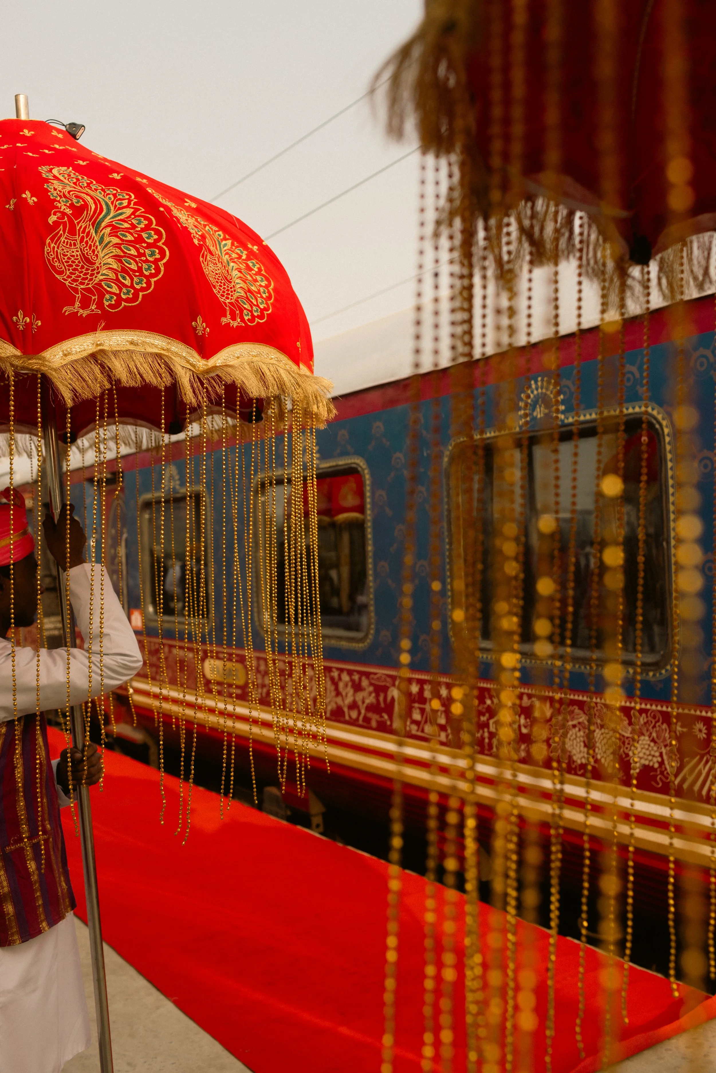 A decorated red umbrella with peacock designs and gold fringe, positioned in front of a decorated styled vehicle with ornate patterns and hanging golden beads.