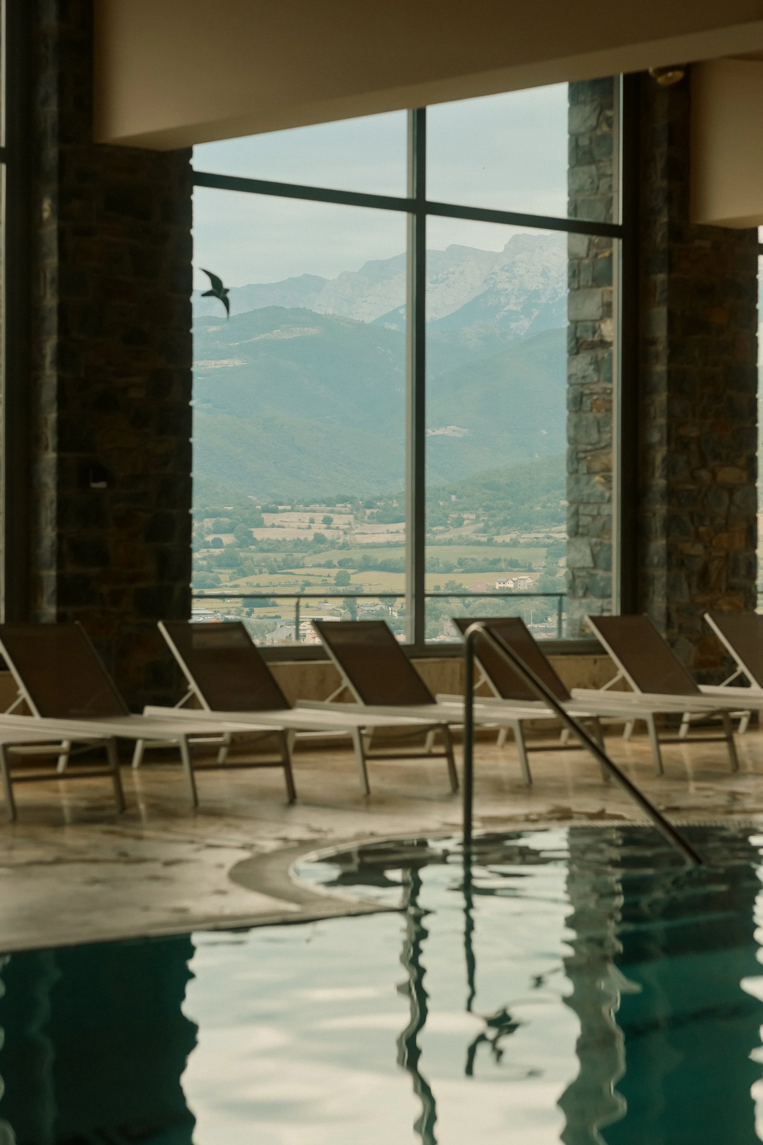 Spa and wellness photography: Indoor pool with lounge chairs and expansive mountain views at El Castell de Ciutat. Professional hospitality portfolio imagery.