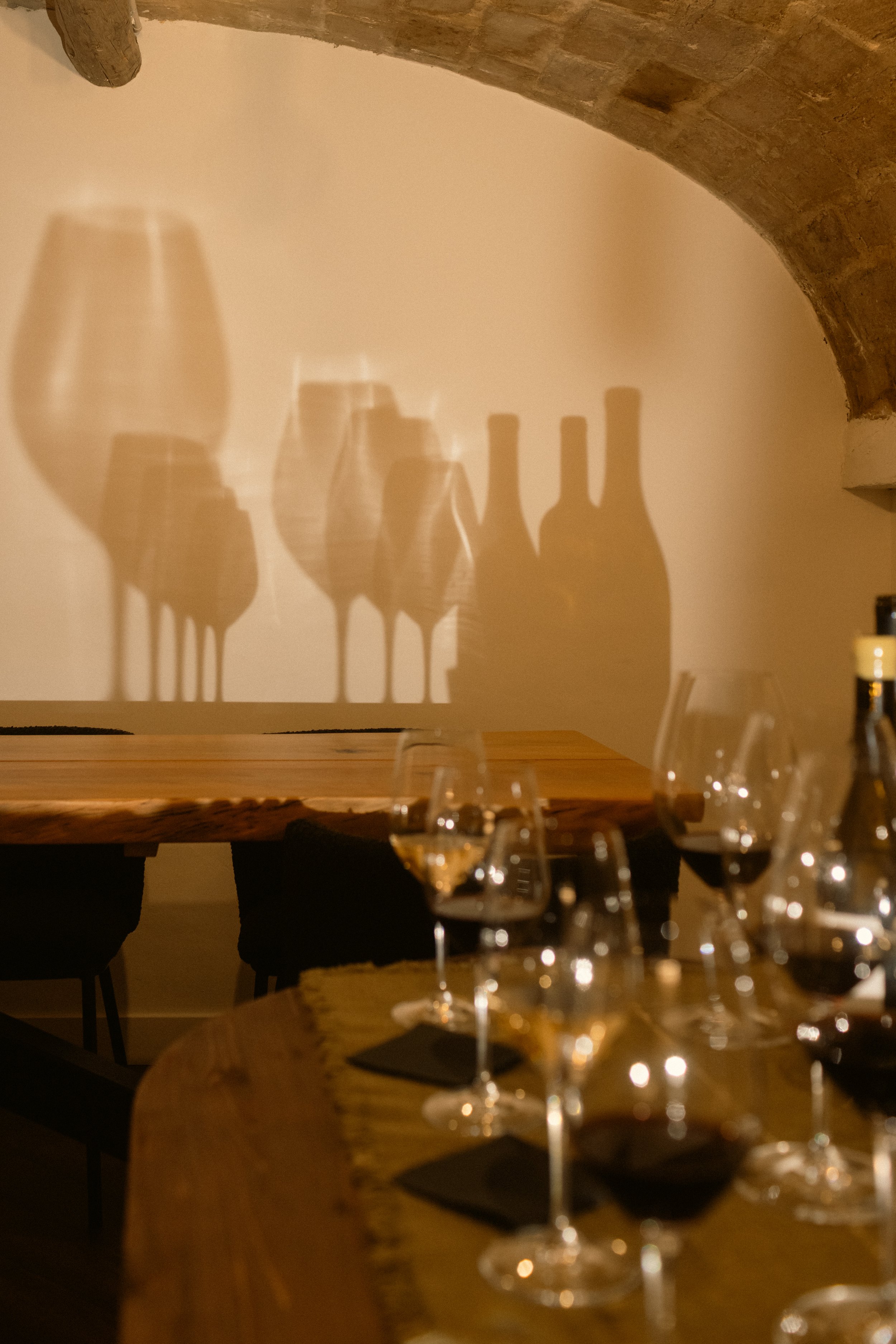 Creative hotel interior photography: dramatic shadows of wine bottles cast on a rustic wall in a cozy dining setting at Ora Priorat. Professional hospitality portfolio imagery.
