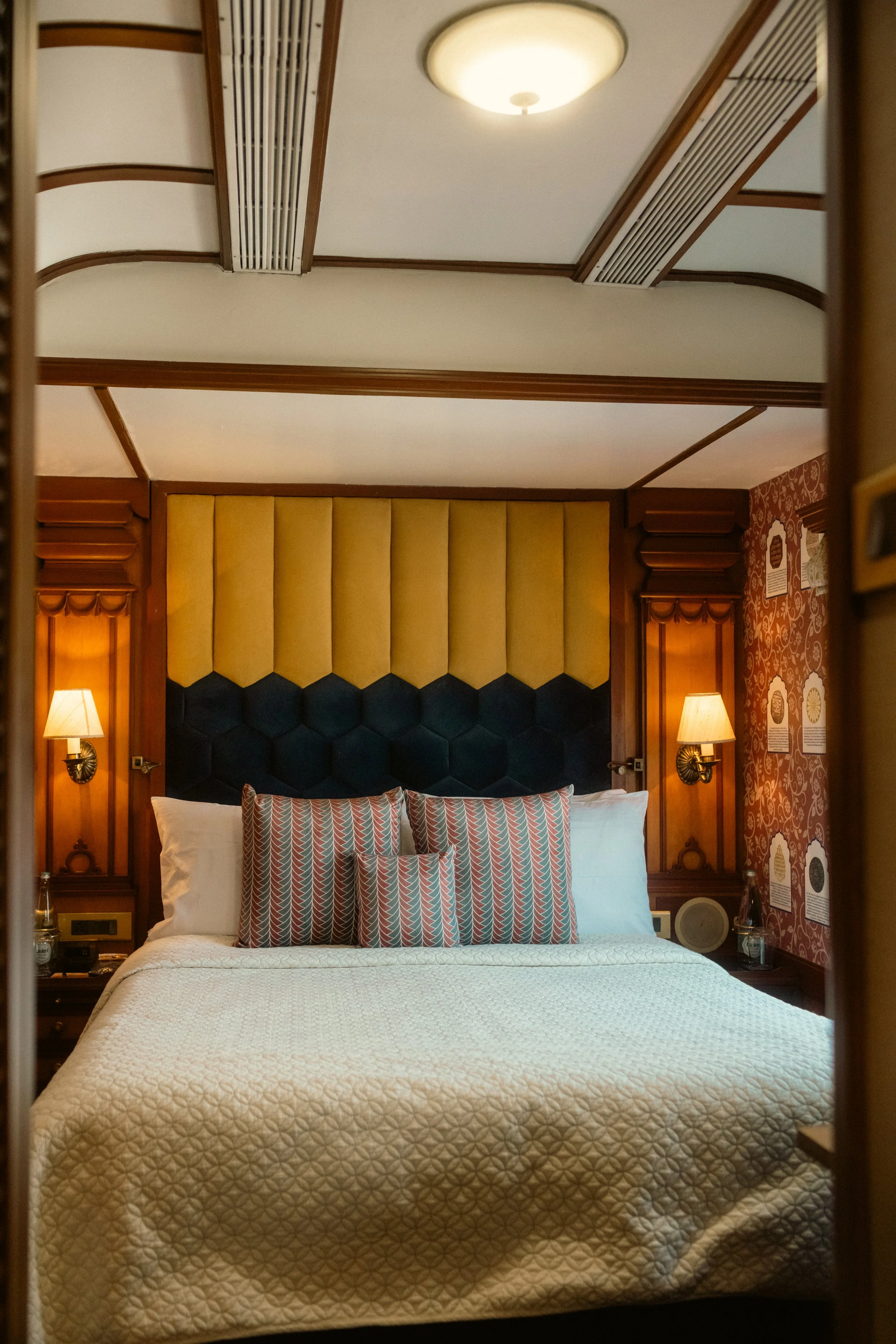 
Luxury hospitality photography: Interior shot of a Deccan Odyssey suite featuring a large padded headboard, ornate wallpaper, and premium bedding. Professional hotel cabin portfolio imagery.