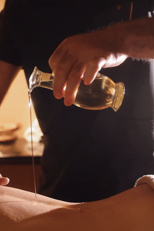 Close-up photograph of hands pouring a thin stream of therapeutic massage oil onto an arm. Relaxation and wellness spa photography.