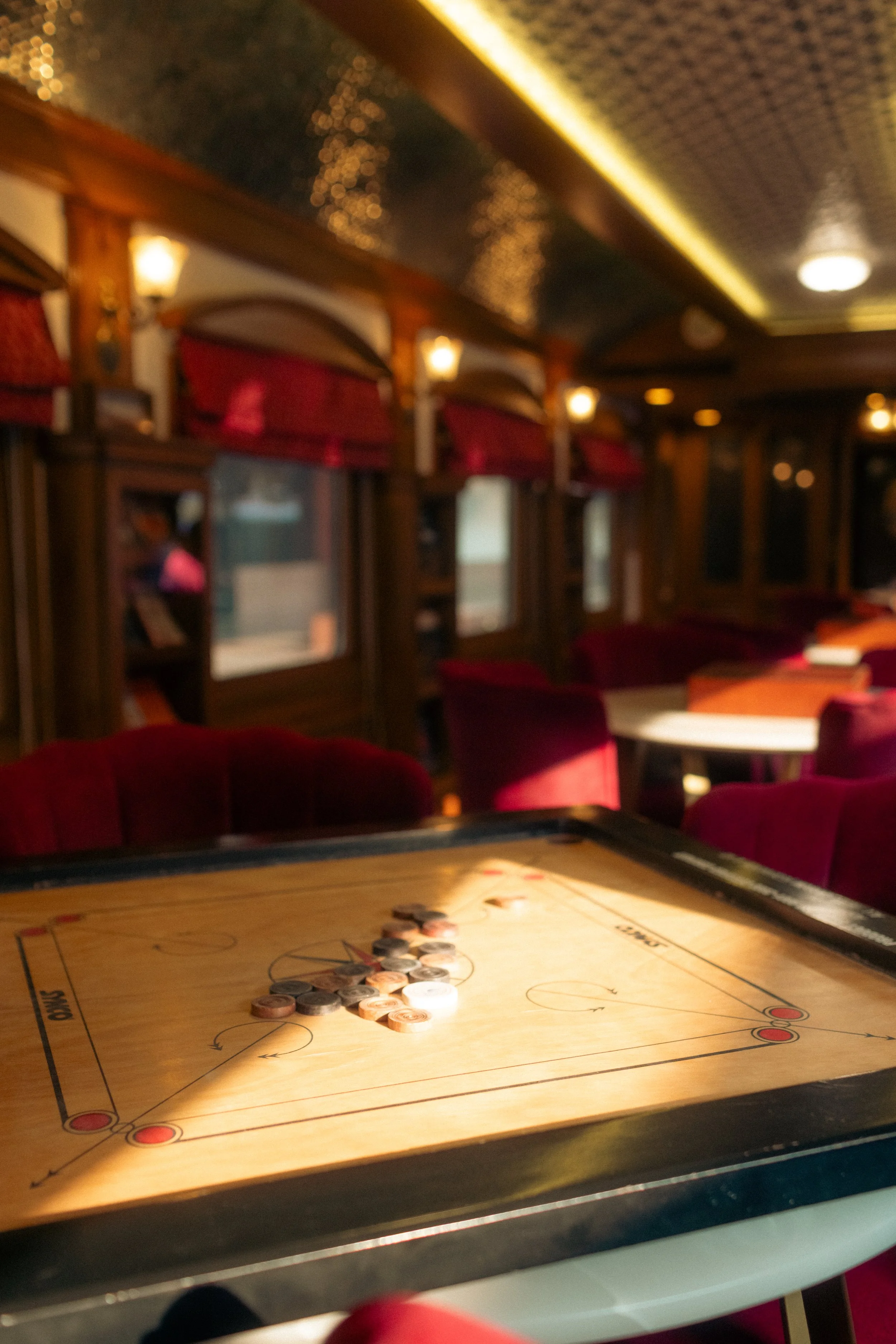 Luxury train interior photography: Vintage carrom board set up in a dimly-lit lounge car with red velvet chairs on the Deccan Odyssey. Professional hospitality and amenitiy portfolio imagery.