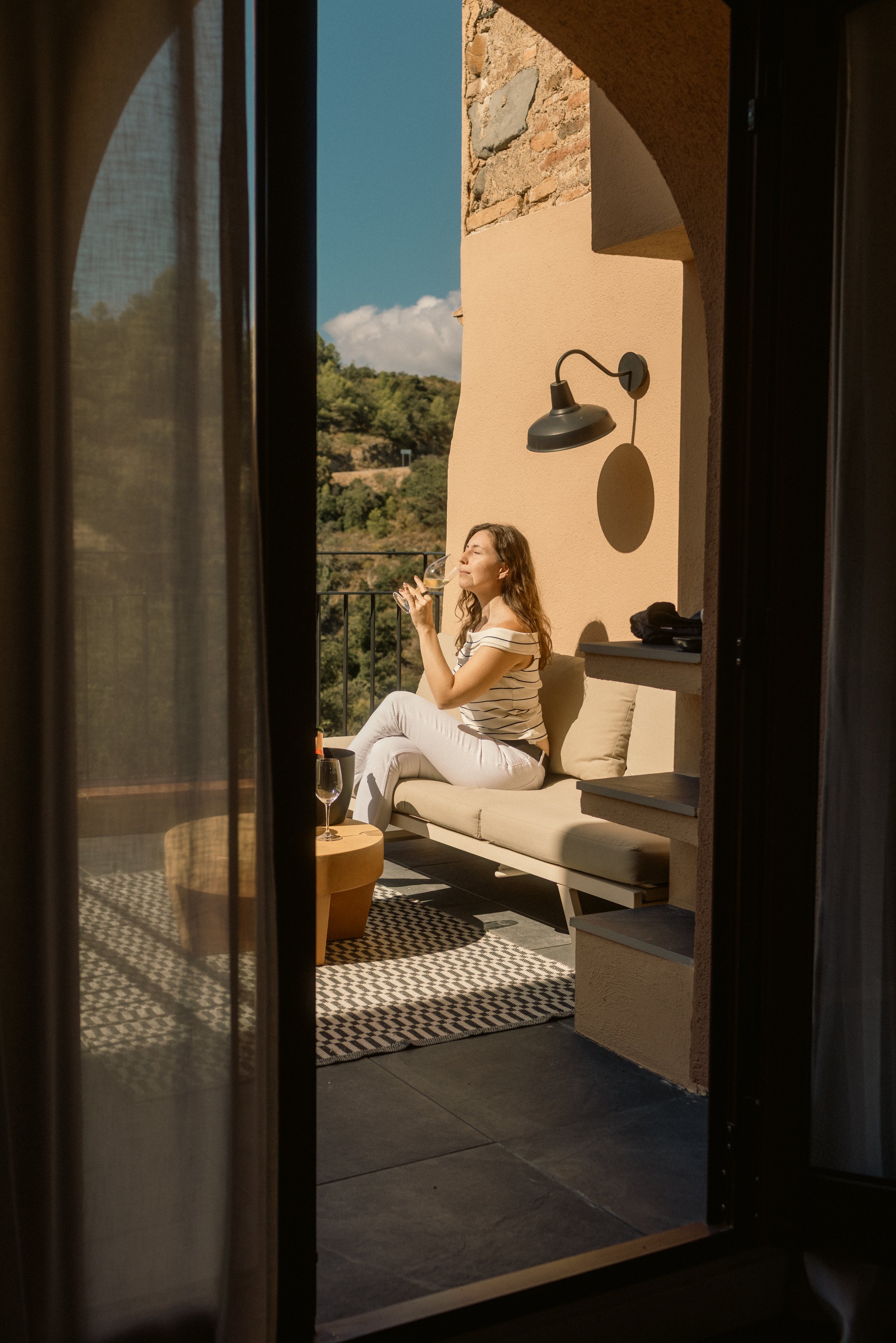 Luxury hotel lifestyle photography: A guest enjoying white wine on a sunny balcony with scenic views. Professional hospitality portfolio shot focusing on outdoor terrace styling at Ora Priorat.