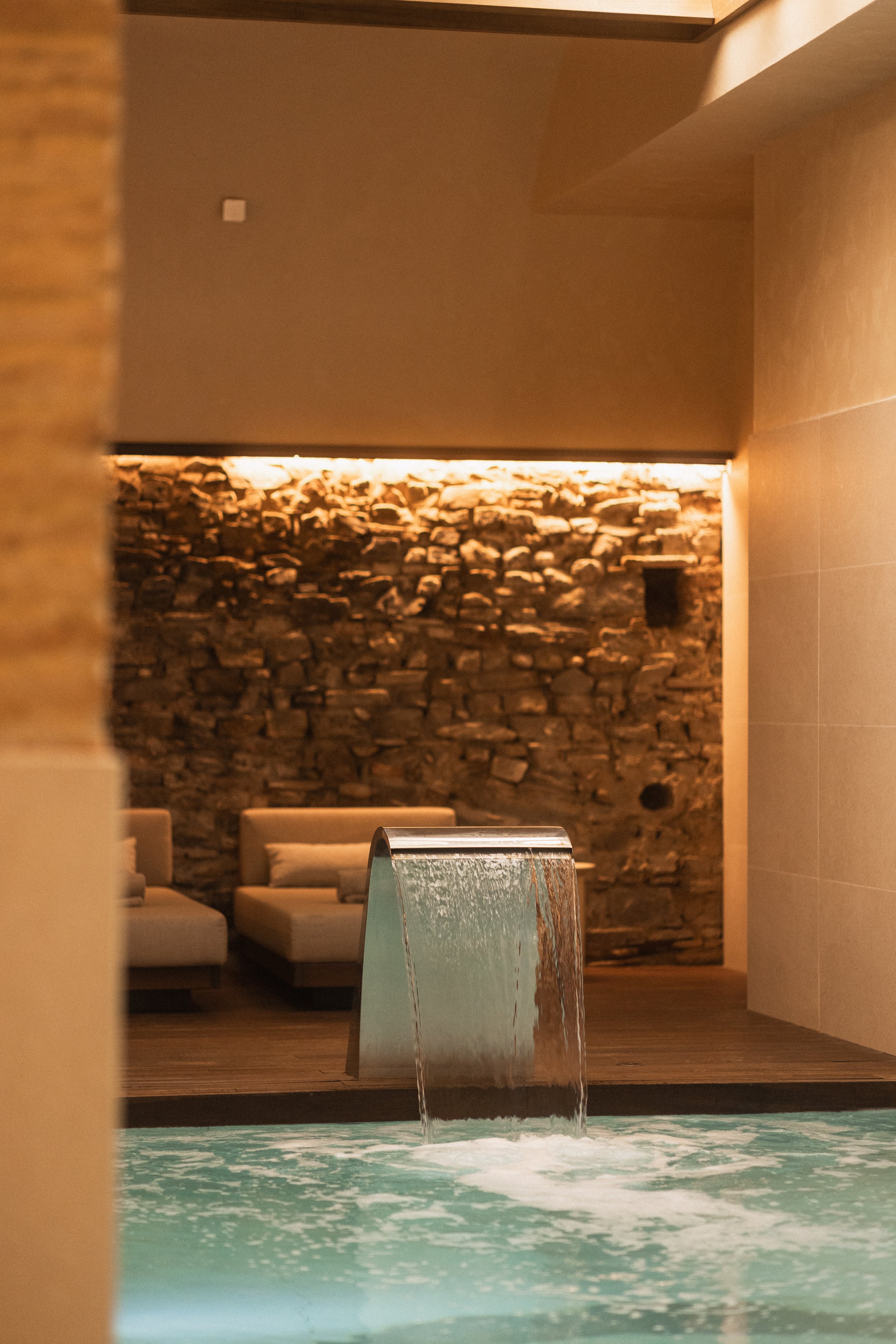 Luxury indoor spa with waterfall feature, rustic stone wall, beige lounge seating, and modern accent lighting. Hotel spa and wellness photography at Mas d’en Bruno.