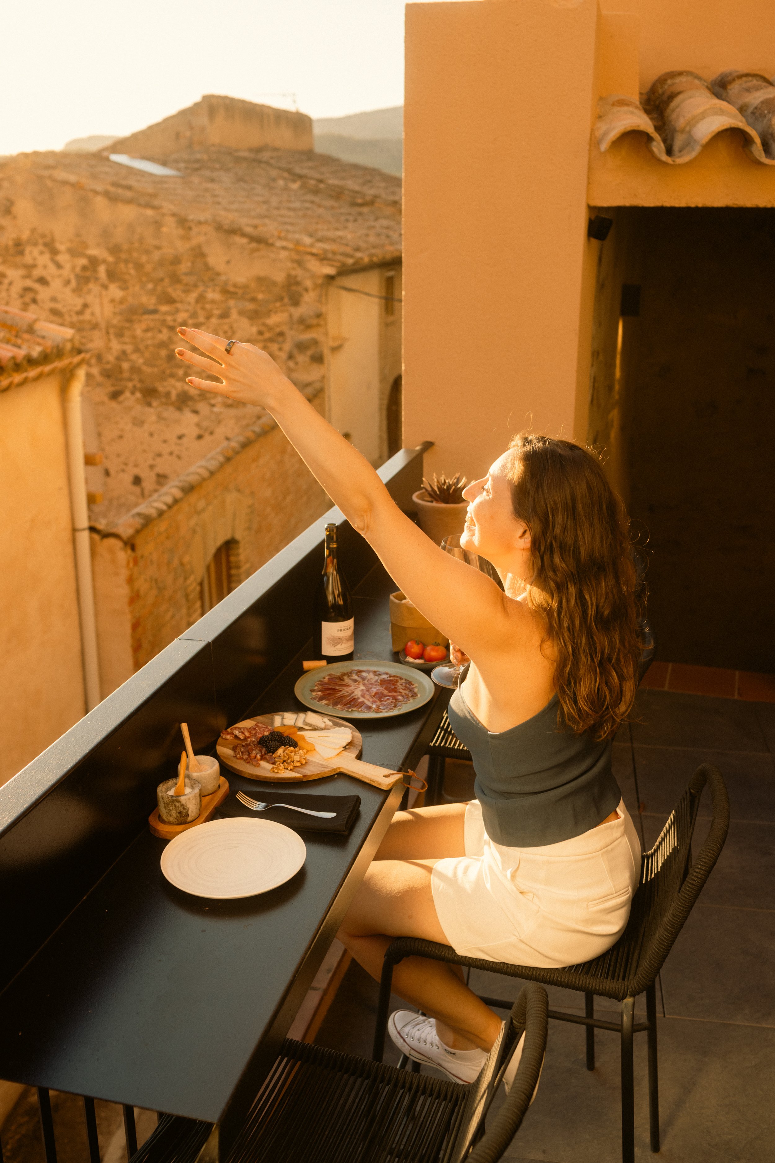 Hotel lifestyle photography: A candid, welcoming shot of a guest on a room balcony with a sunset view and cheese platter, reaching out to someone below. Example of natural light hospitality imagery at Ora Priorat.