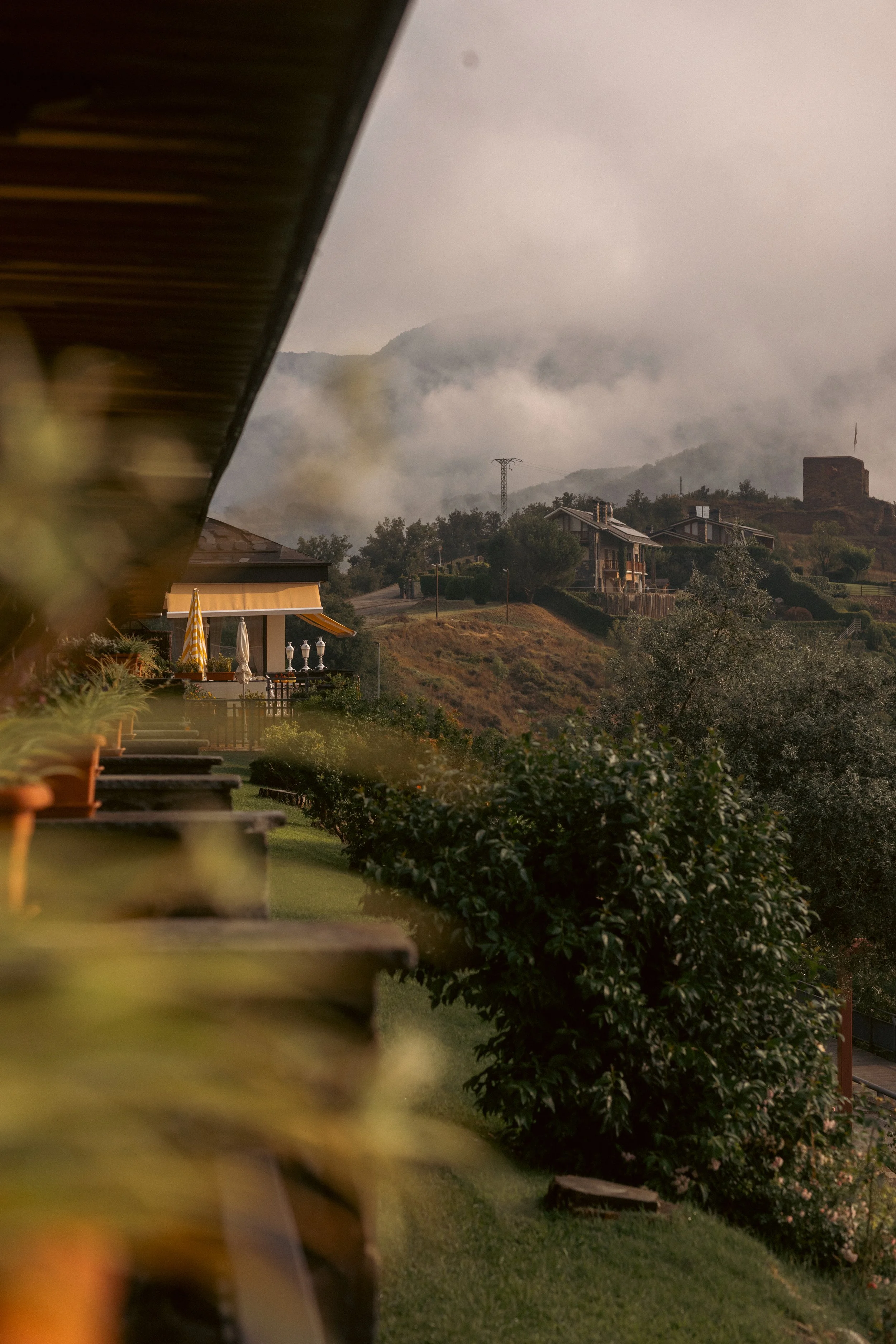 Architectural and landscape photography: A moody, scenic shot of a hillside neighborhood shrouded in fog, viewed from a hotel balcony at El Castell de Ciutat. Professional hospitality and location imagery.