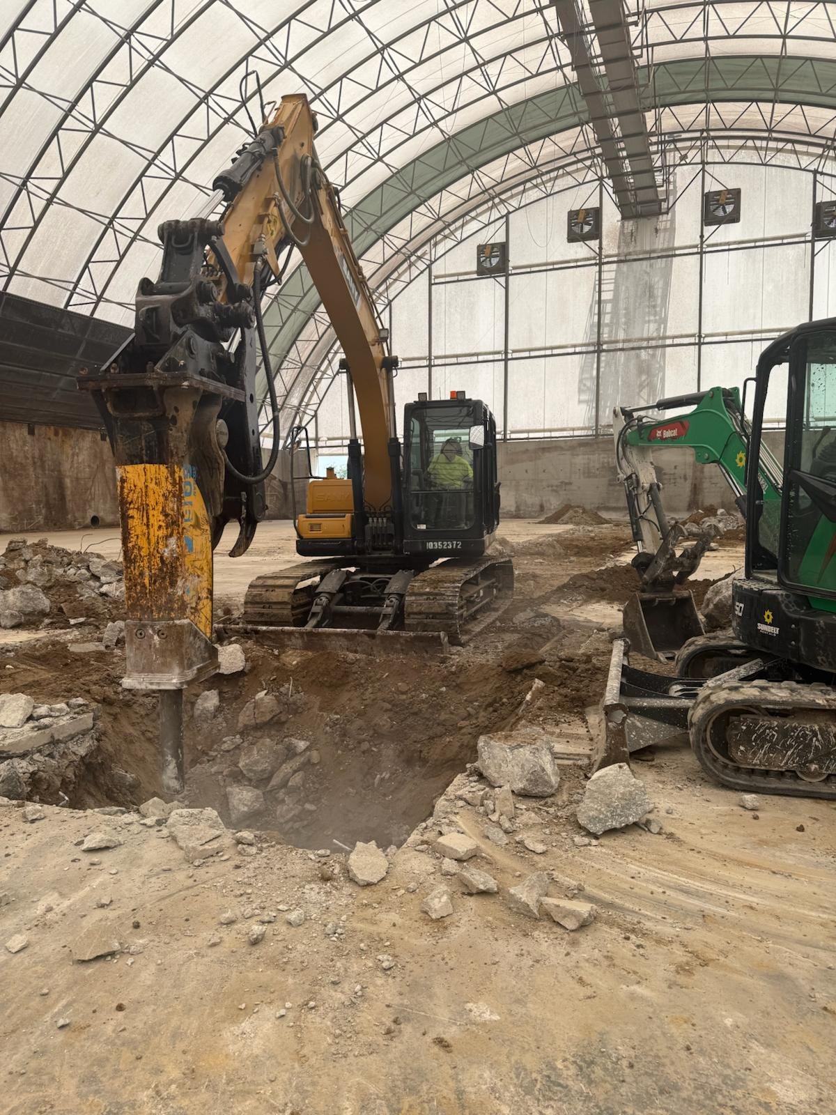 Construction site inside a large arched building with two excavators digging a foundation or trench.