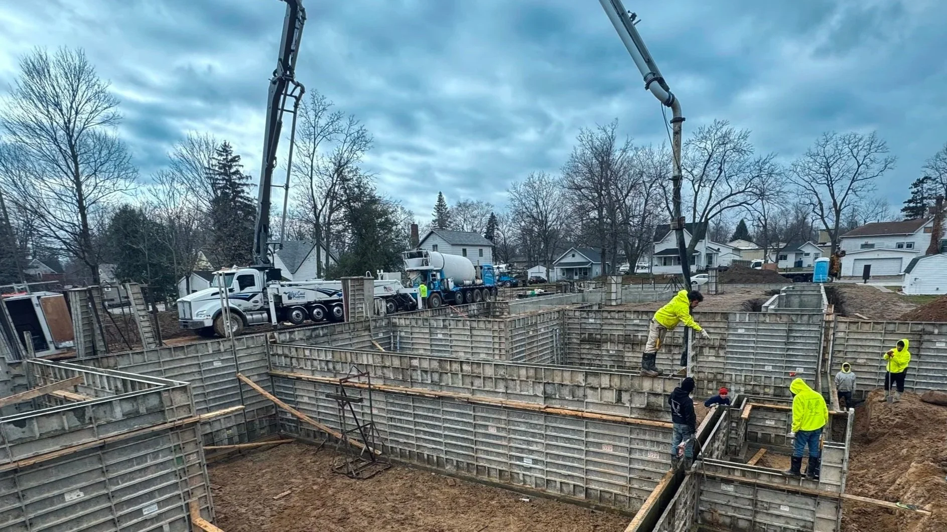 Construction workers are working on the foundation of a building, using concrete pumps and formwork, in a residential neighborhood with houses and trees in the background.