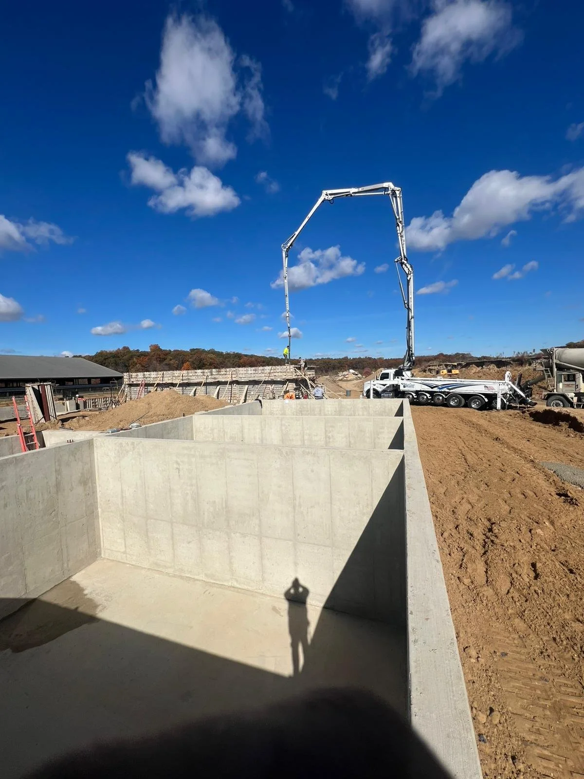 Construction site with large cement walls, a concrete pump truck, and a person standing on the top of the structure, under a blue sky with clouds.