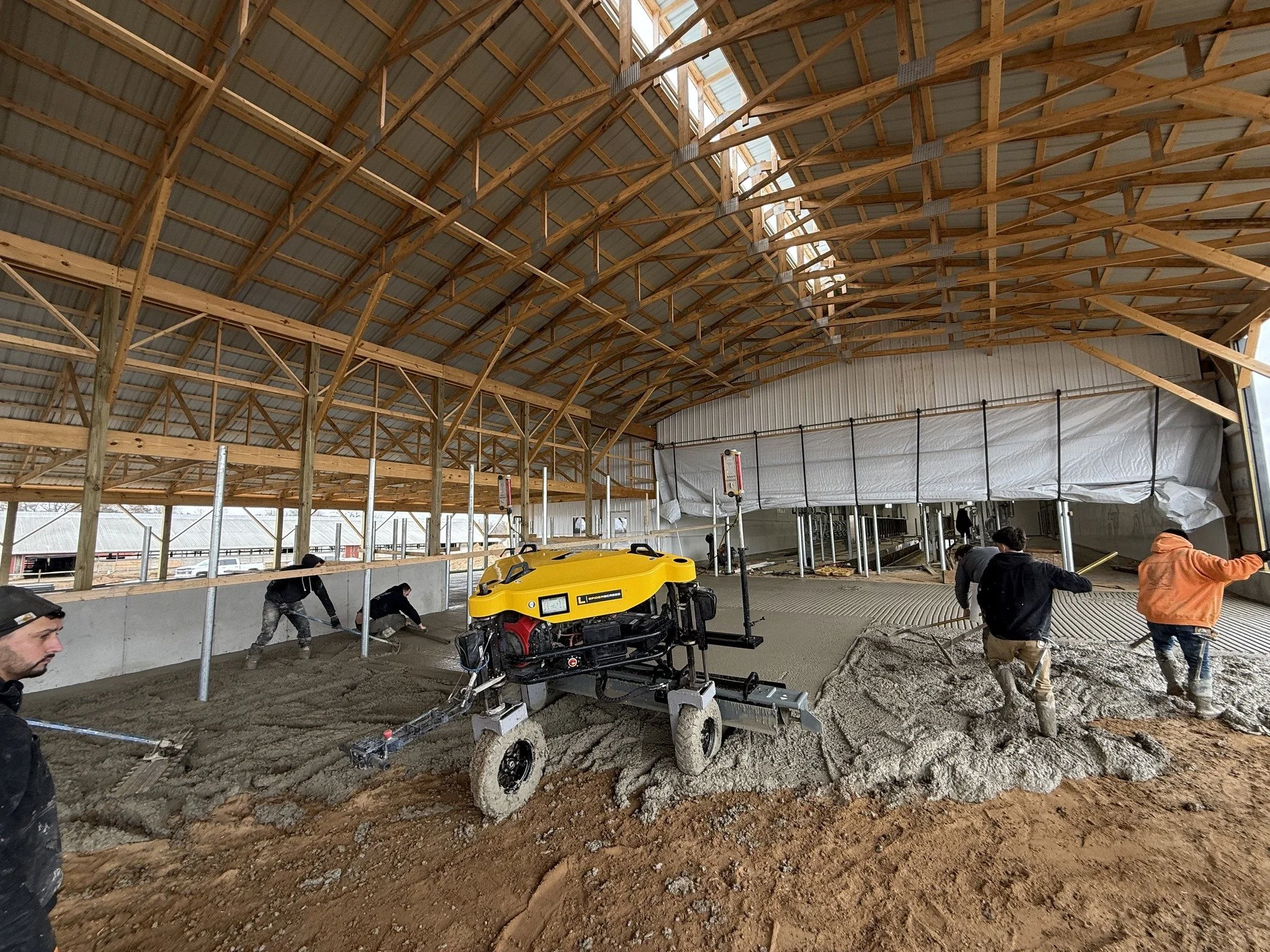 Construction workers are pouring and leveling concrete inside a large wooden-framed building with a high curved ceiling.