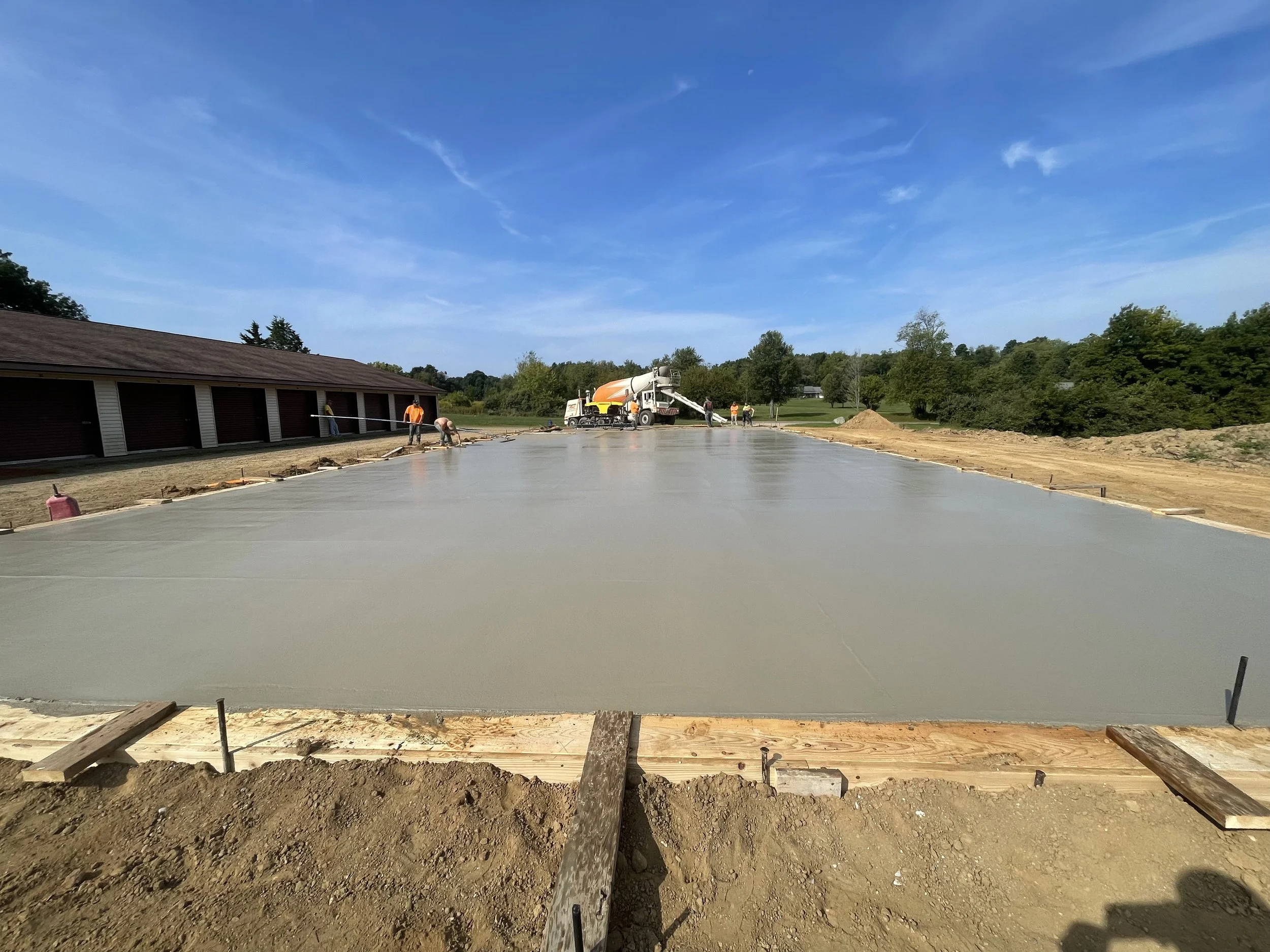 Construction workers pour concrete onto a foundation slab at a building site on a sunny day with blue sky and trees in the background.