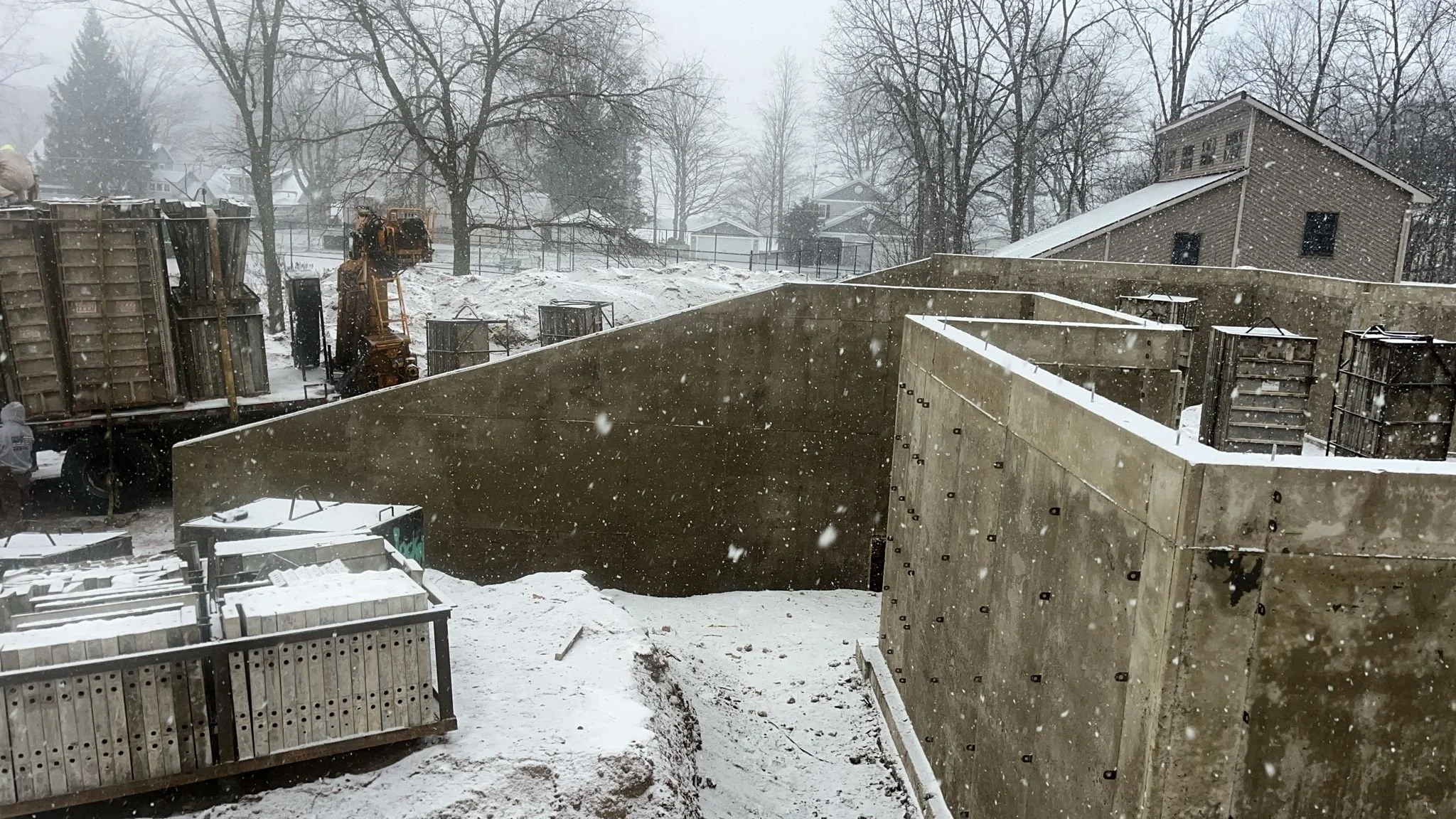 Construction site in snowy weather with large concrete walls, construction materials, and a truck with equipment. Snow is falling, and leafless trees and houses are in the background.