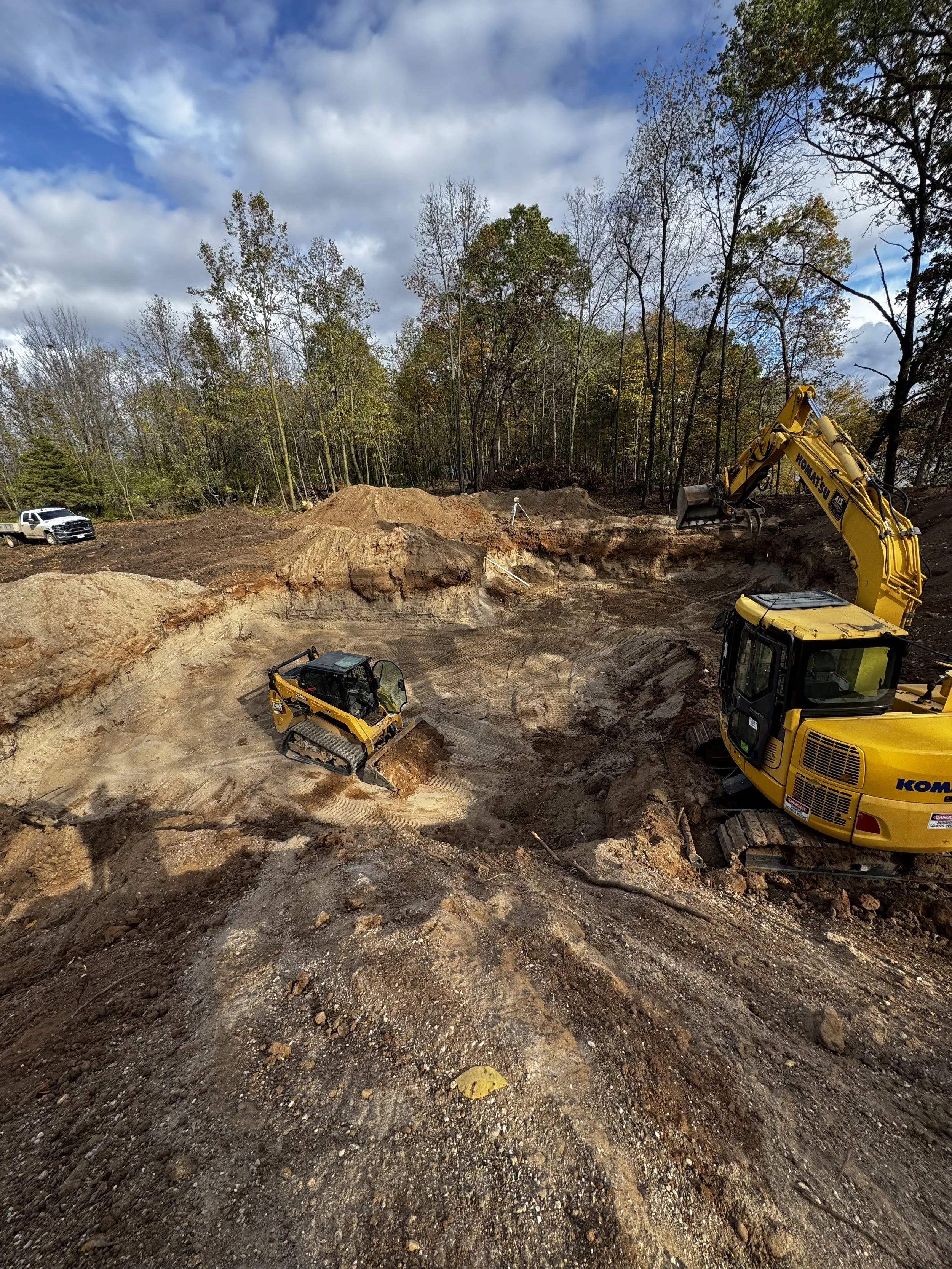 Construction site with a yellow excavator and a small tracked loader working on dirt and excavation, surrounded by trees under a partly cloudy sky.