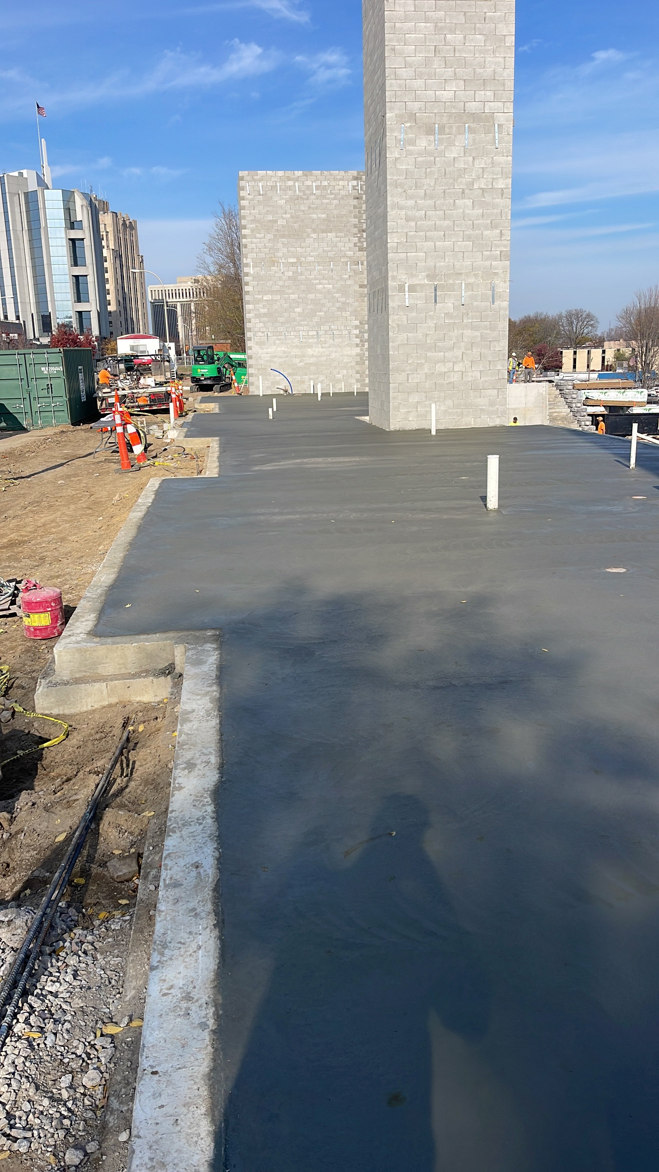 Construction site with fresh concrete pavement, several white pipe markers, and workers in the background near a gray brick tower structure.