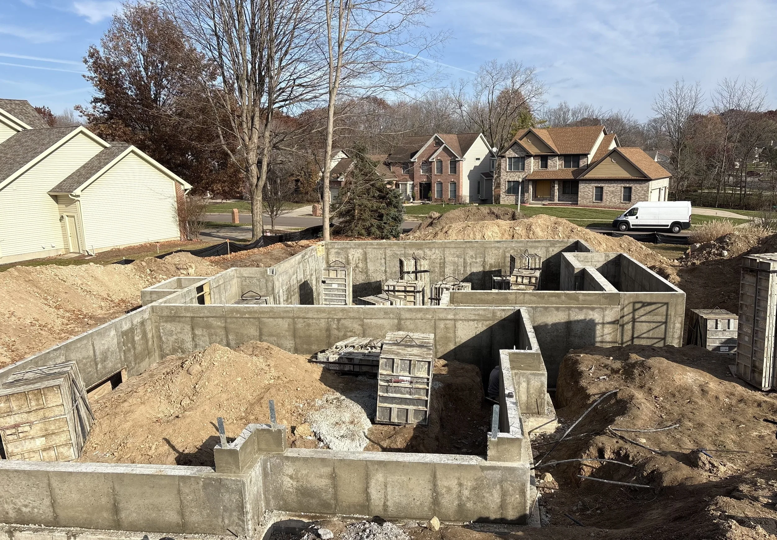 Construction site with concrete foundation and dirt mounds, residential neighborhood with houses and trees in the background, a white van parked on the street.
