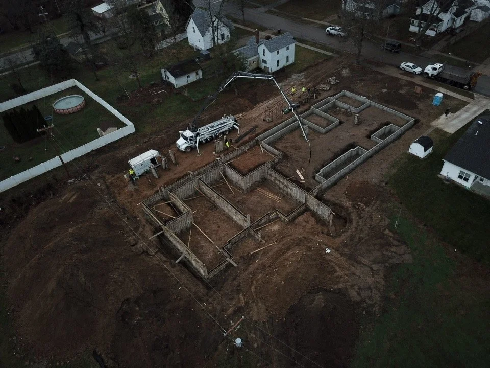 Aerial view of a residential construction site with foundation walls and concrete pouring in progress, surrounded by neighboring houses, vehicles, and a fenced backyard.