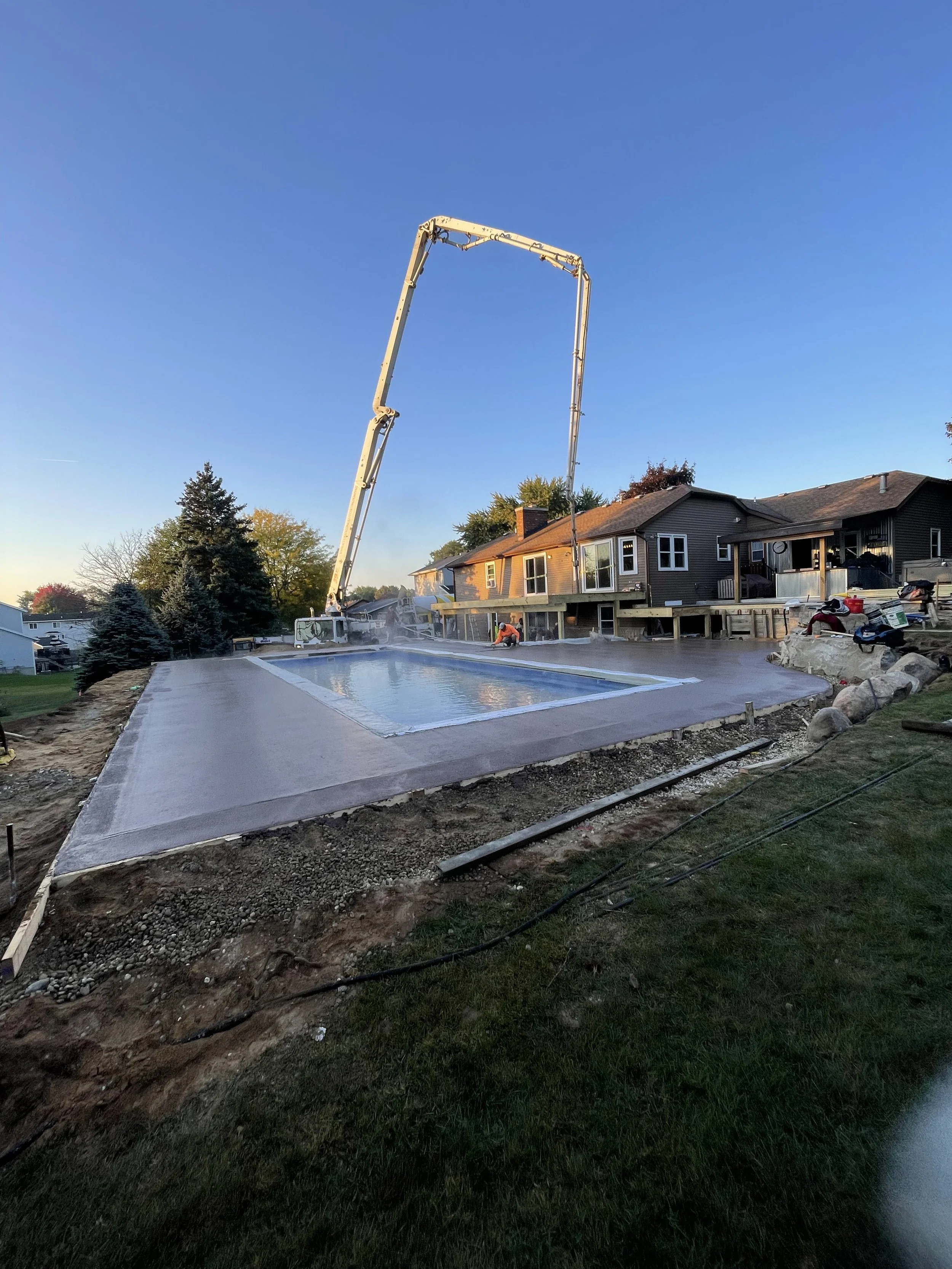 Construction workers pour concrete into a pool at a residential backyard, with houses and trees in the background.