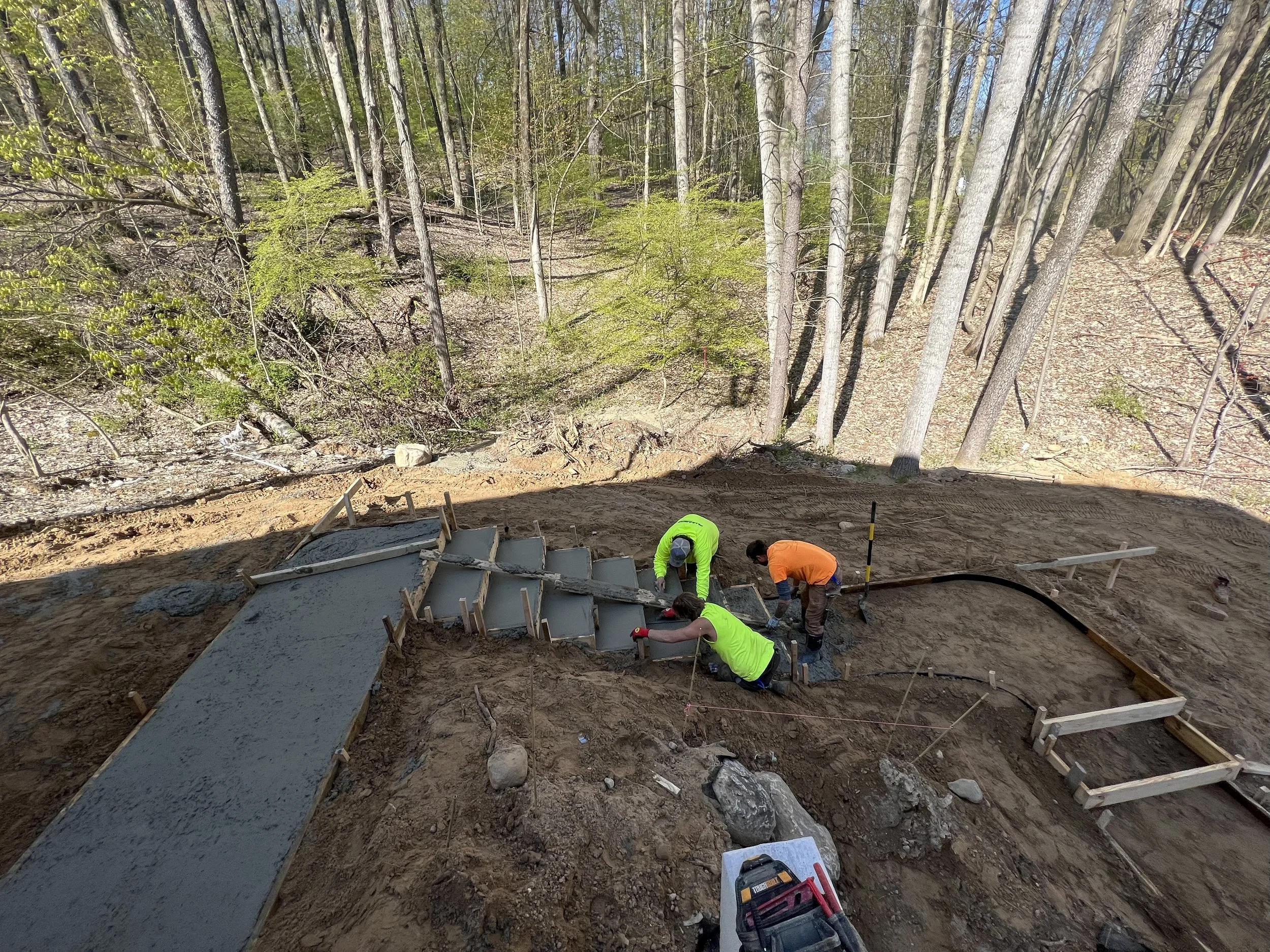 Workers pouring and smoothing concrete for a staircase on a construction site in a forested area.