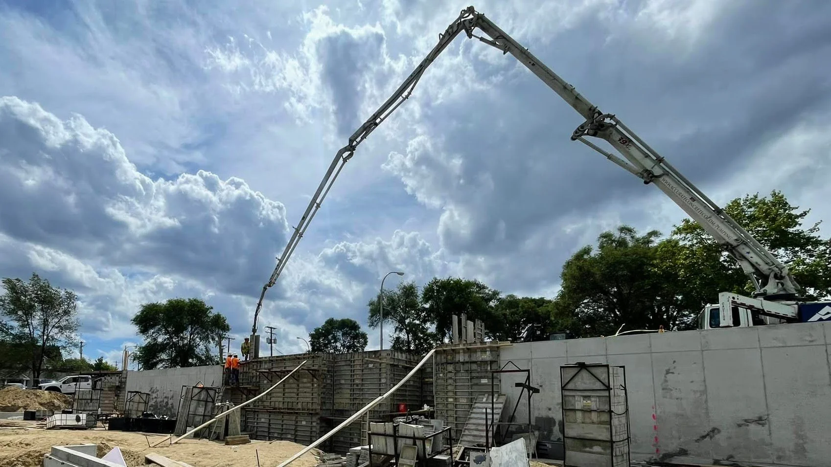 Construction workers and equipment working on a concrete wall with a large crane arm extending overhead, set against cloudy sky.