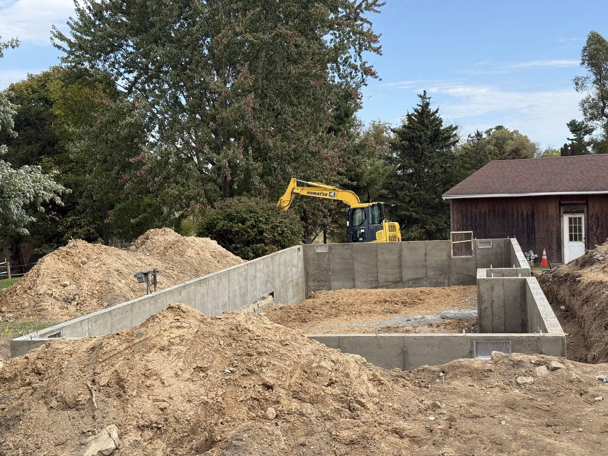 Construction site with a small yellow excavator working near concrete foundations, piles of dirt, and surrounded by trees and a small wooden building with a door and traffic cone.