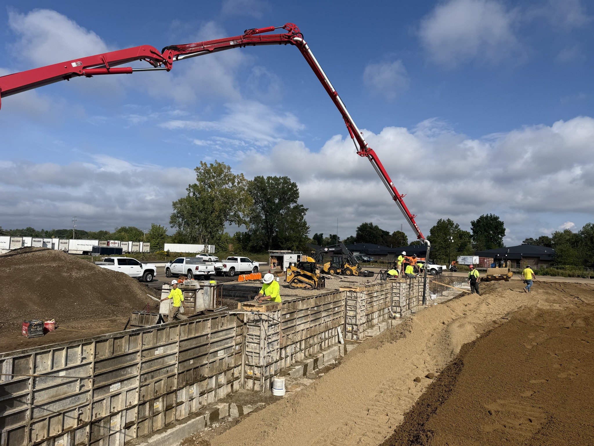 Construction site with workers in high-visibility yellow clothing building a concrete wall, large red concrete pump extending over the site, and dirt piles on the ground, under a partly cloudy sky.