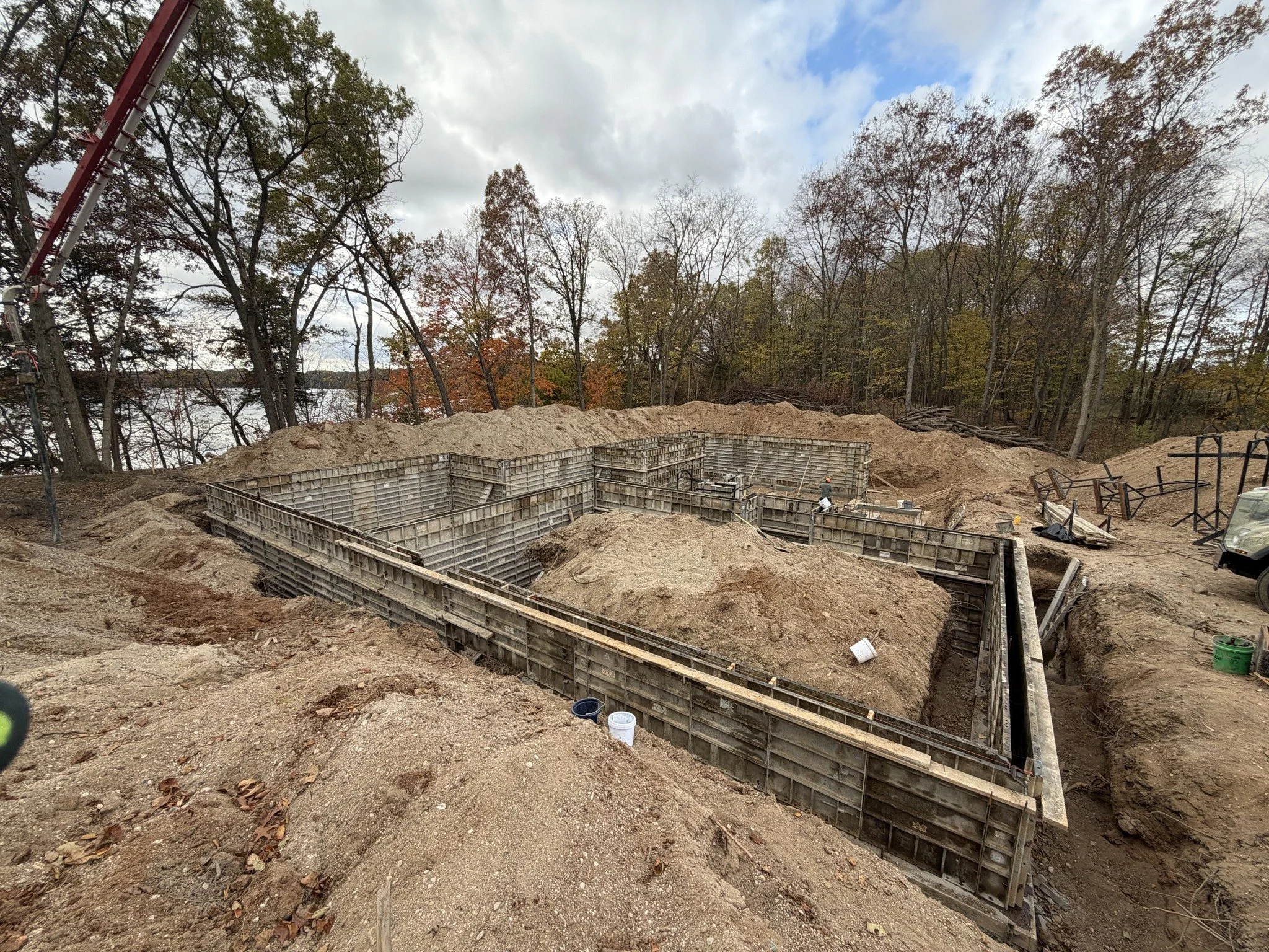 Construction site with concrete foundation forms, dirt, and construction workers near a wooded area with fall foliage.