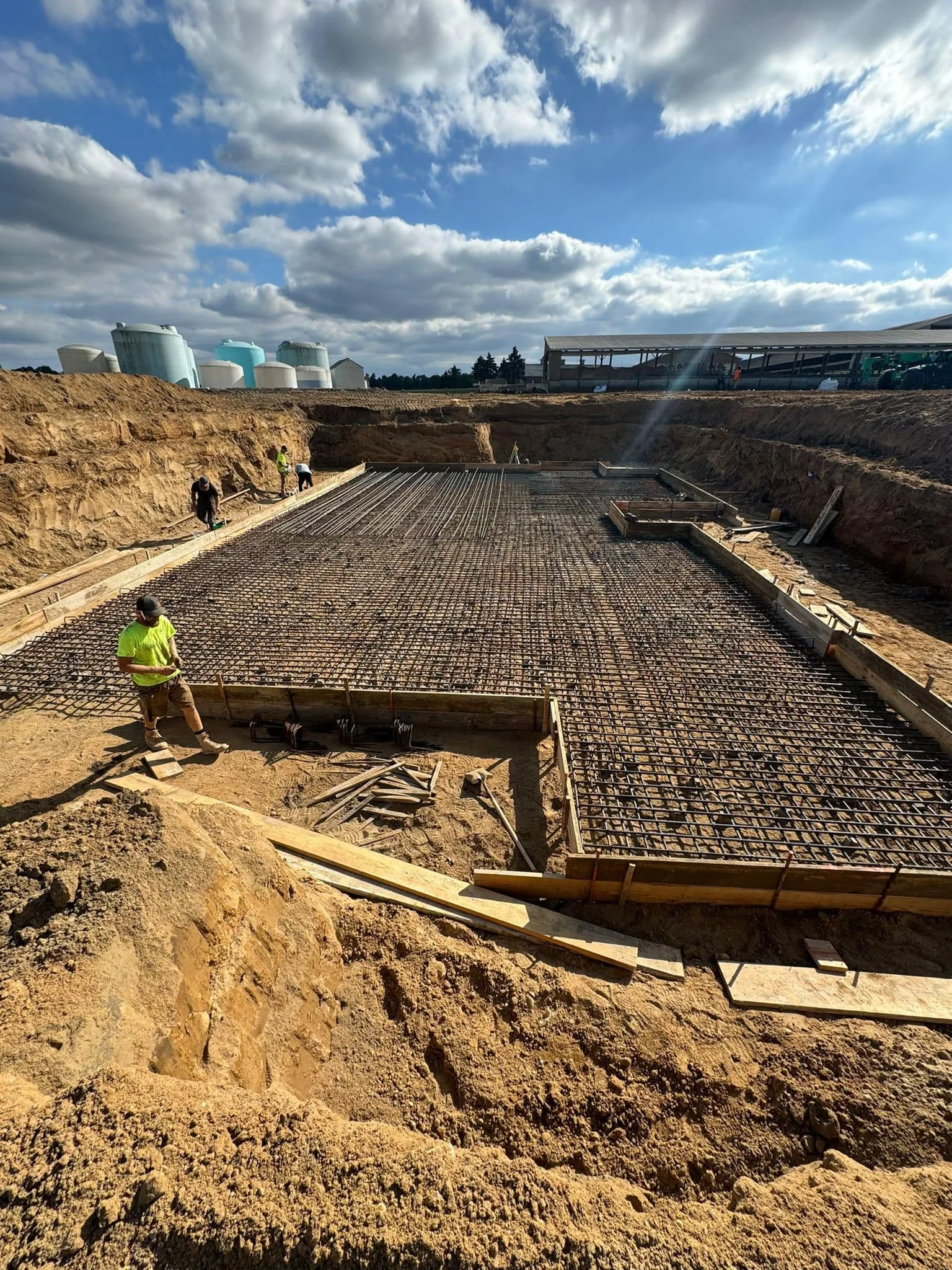 Construction site with workers building a foundation, reinforced with steel rebar, under a partly cloudy sky.
