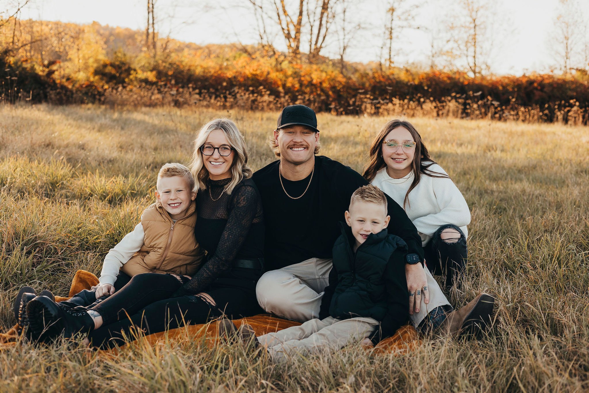 Family of six sitting on a blanket in a grassy field during autumn with trees and hills in the background, smiling at the camera.