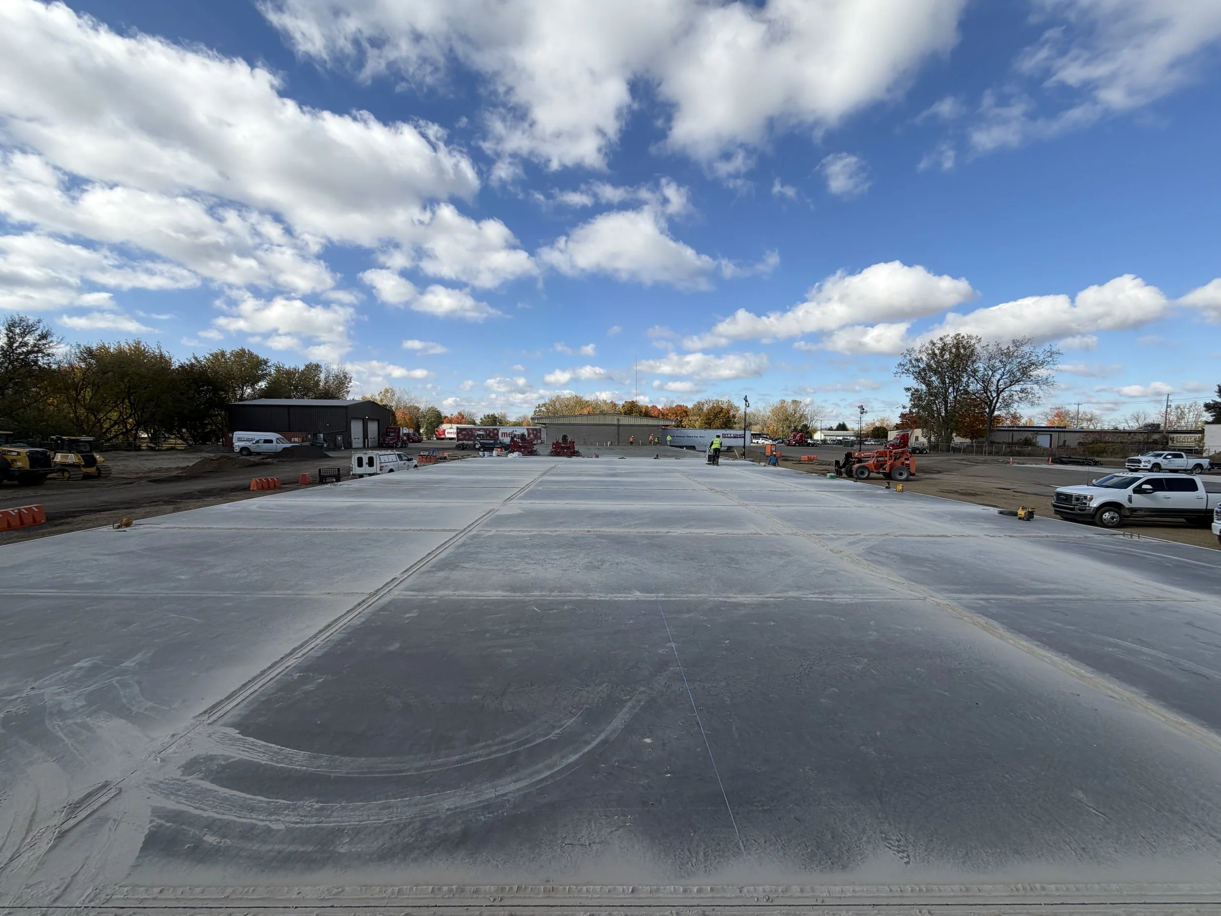 Construction site with a large flat surface, construction vehicles, and trucks parked in the background under a partly cloudy sky.