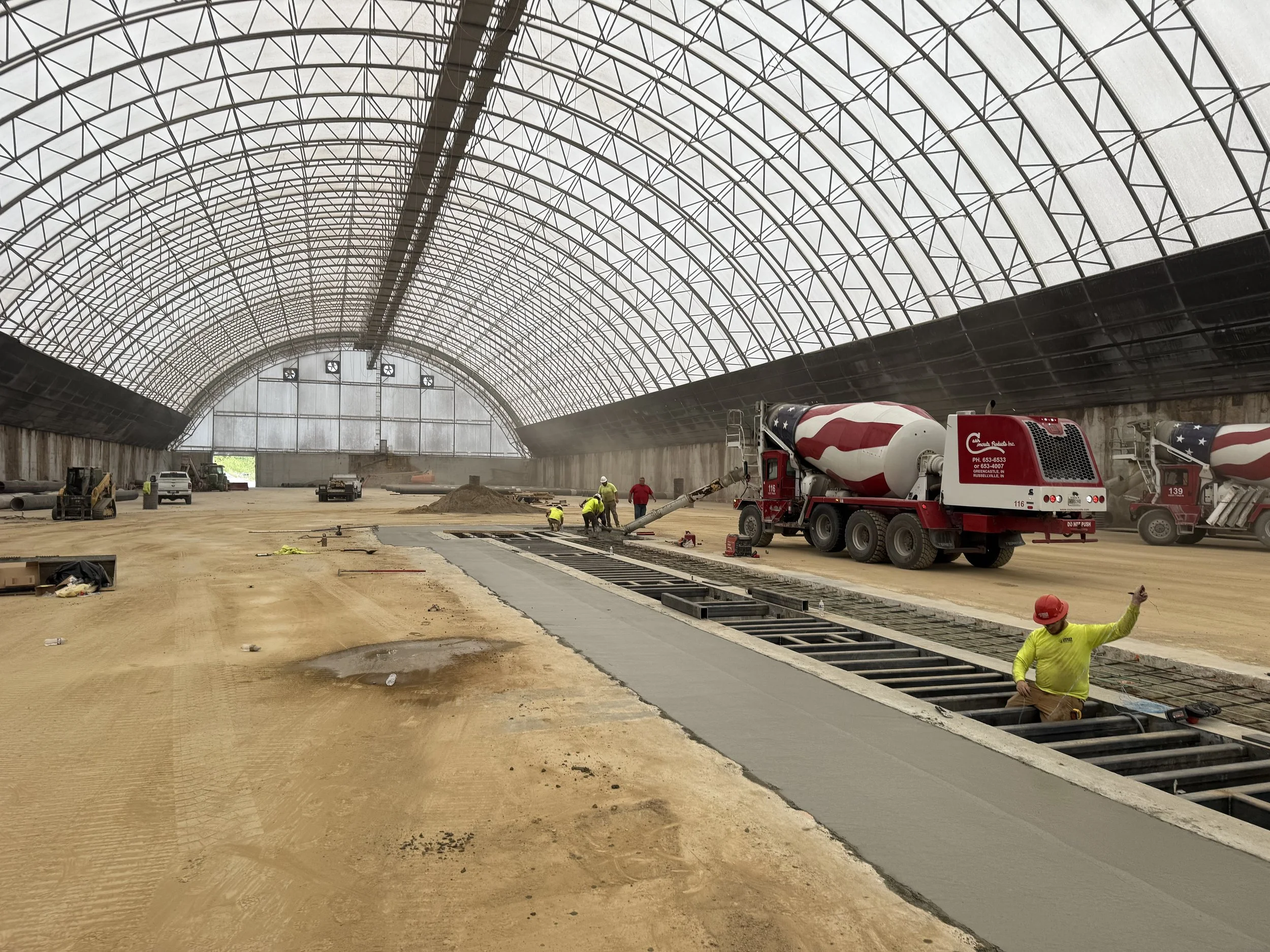 Construction workers inside a large covered building with an arched ceiling, pouring concrete on the ground, using cement mixers and construction equipment.