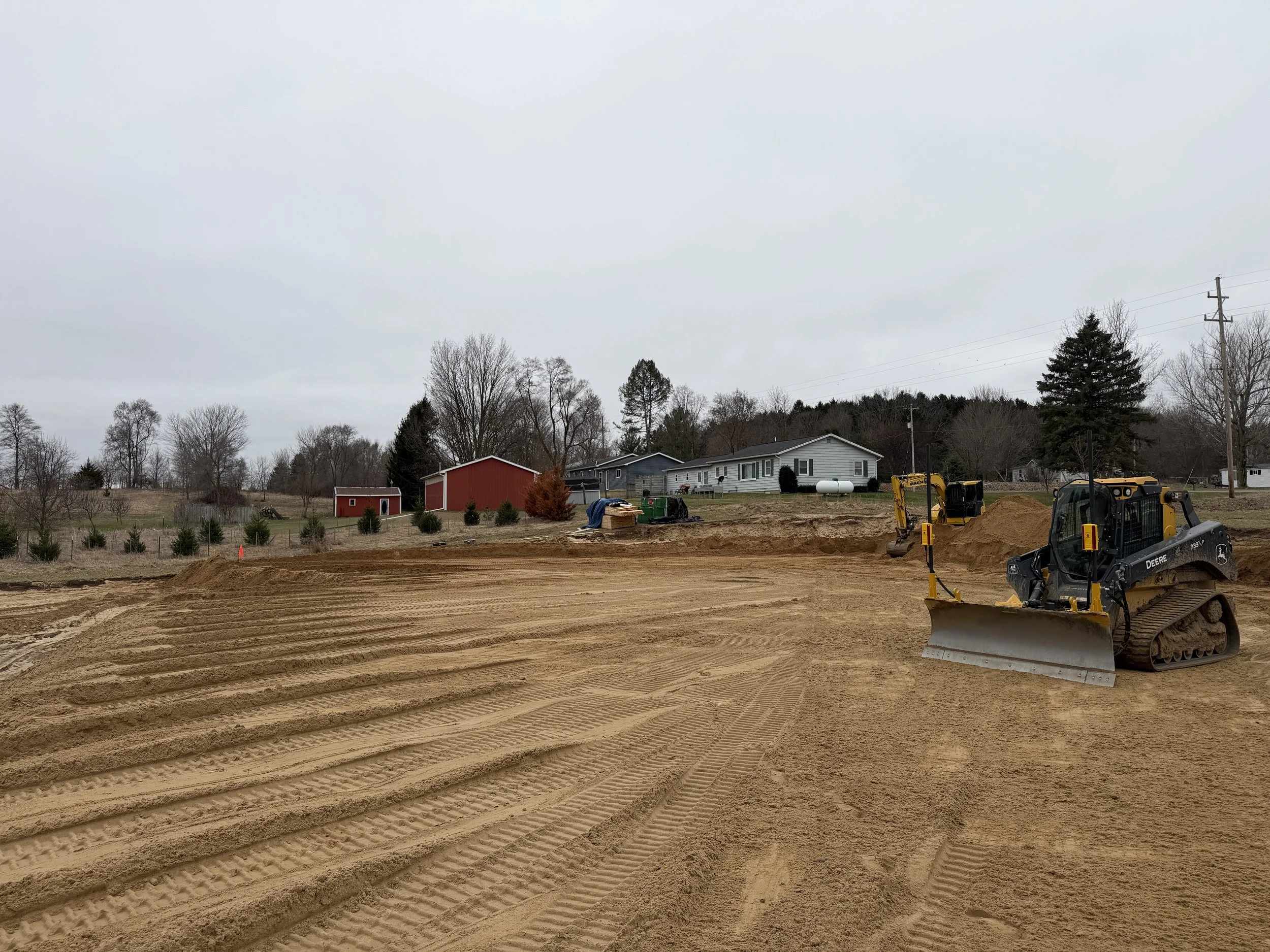 Construction site with a dirt-covered ground, a small black and yellow bulldozer, and a yellow excavator in the background, with residential houses and cloudy sky in the distance.