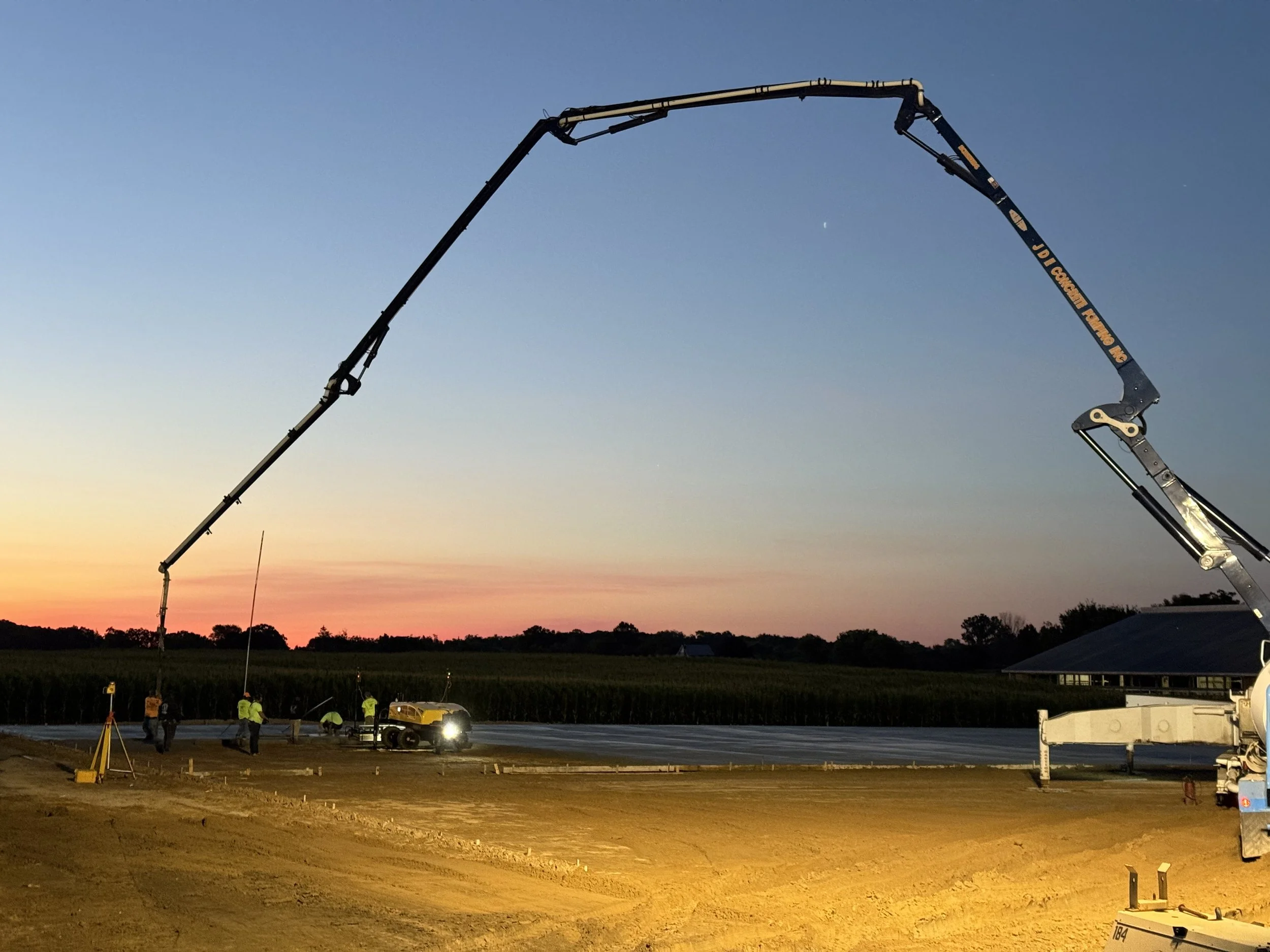 Construction workers laying asphalt on a road at sunset, with a large concrete pump truck and a roller in use.