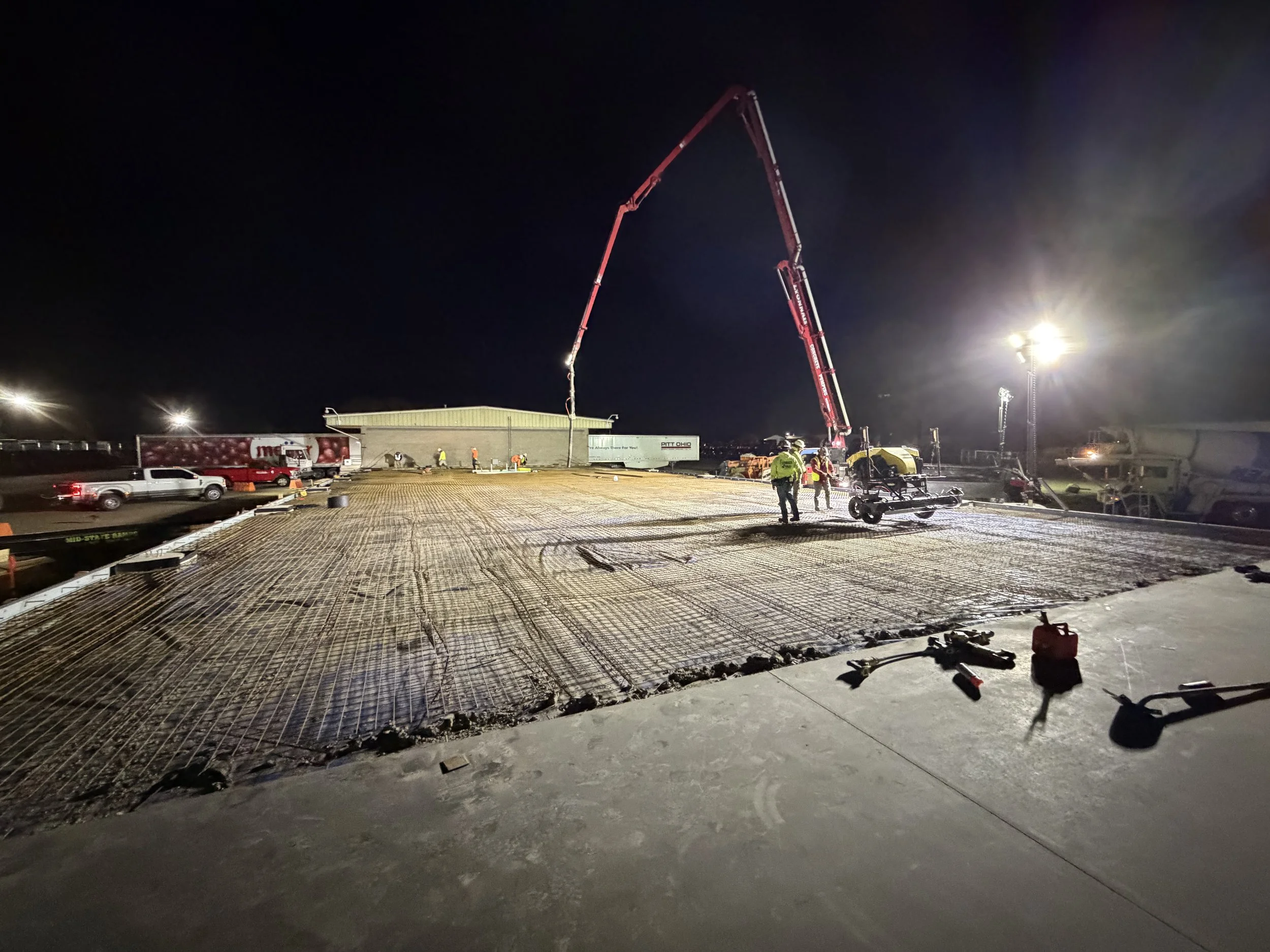 Nighttime construction site with workers, concrete pouring equipment, and bright lights illuminating the area.