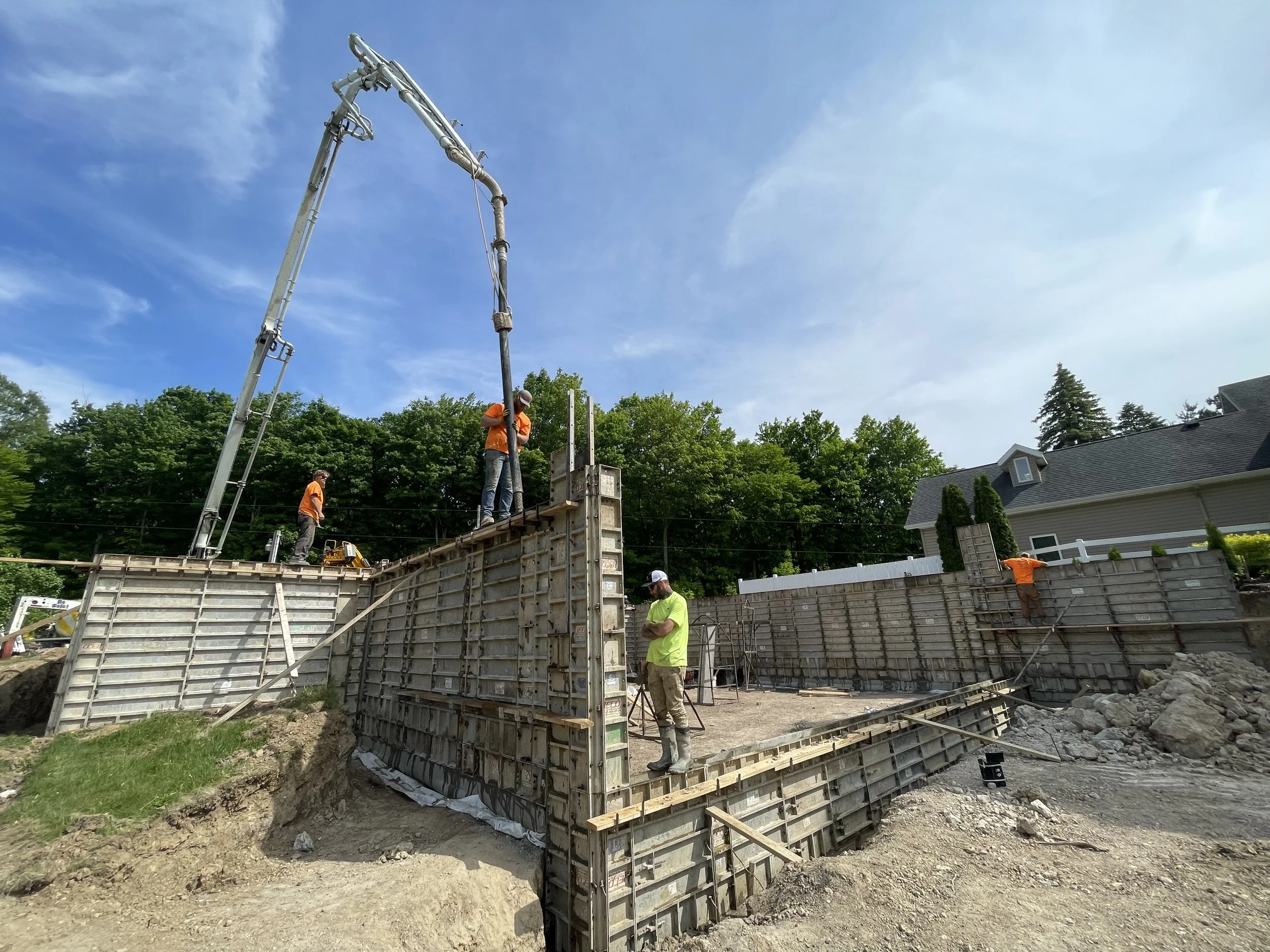 Construction workers building a foundation with concrete forms at a residential site, using a concrete pumping truck and surrounded by trees and a house in the background.