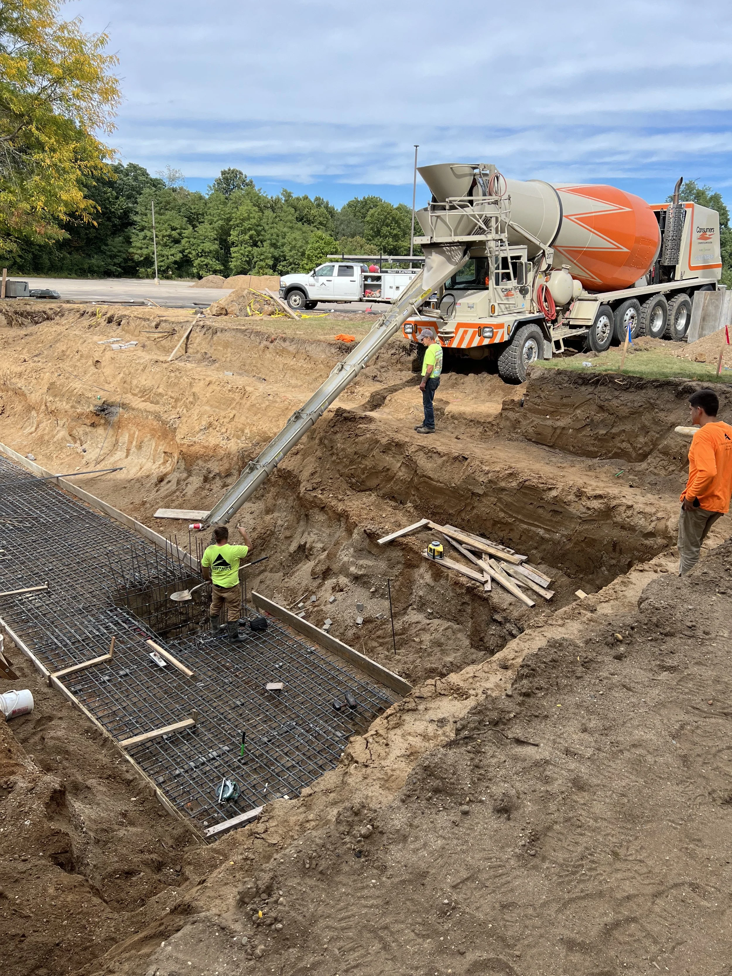 Construction site with workers pouring concrete into a foundation, using a concrete mixer truck and chute, with rebar reinforcement in place.
