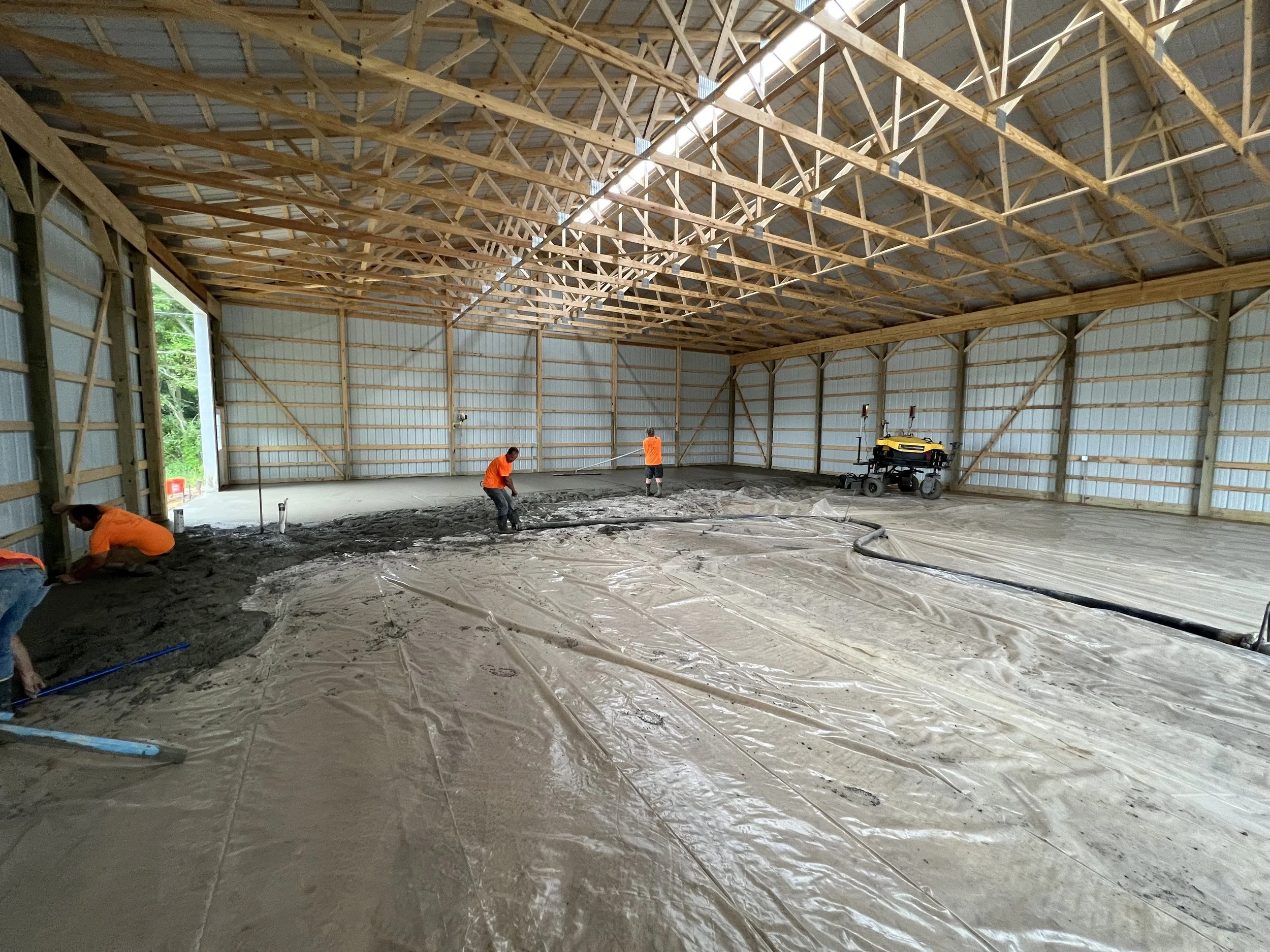 Workers in orange shirts pouring and leveling concrete inside a large, open steel-frame building under construction.