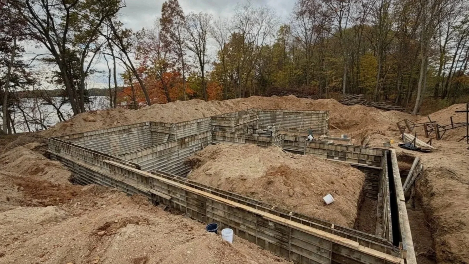 Construction site with excavated foundation and wooden formwork for a building, surrounded by dirt and trees with autumn foliage.