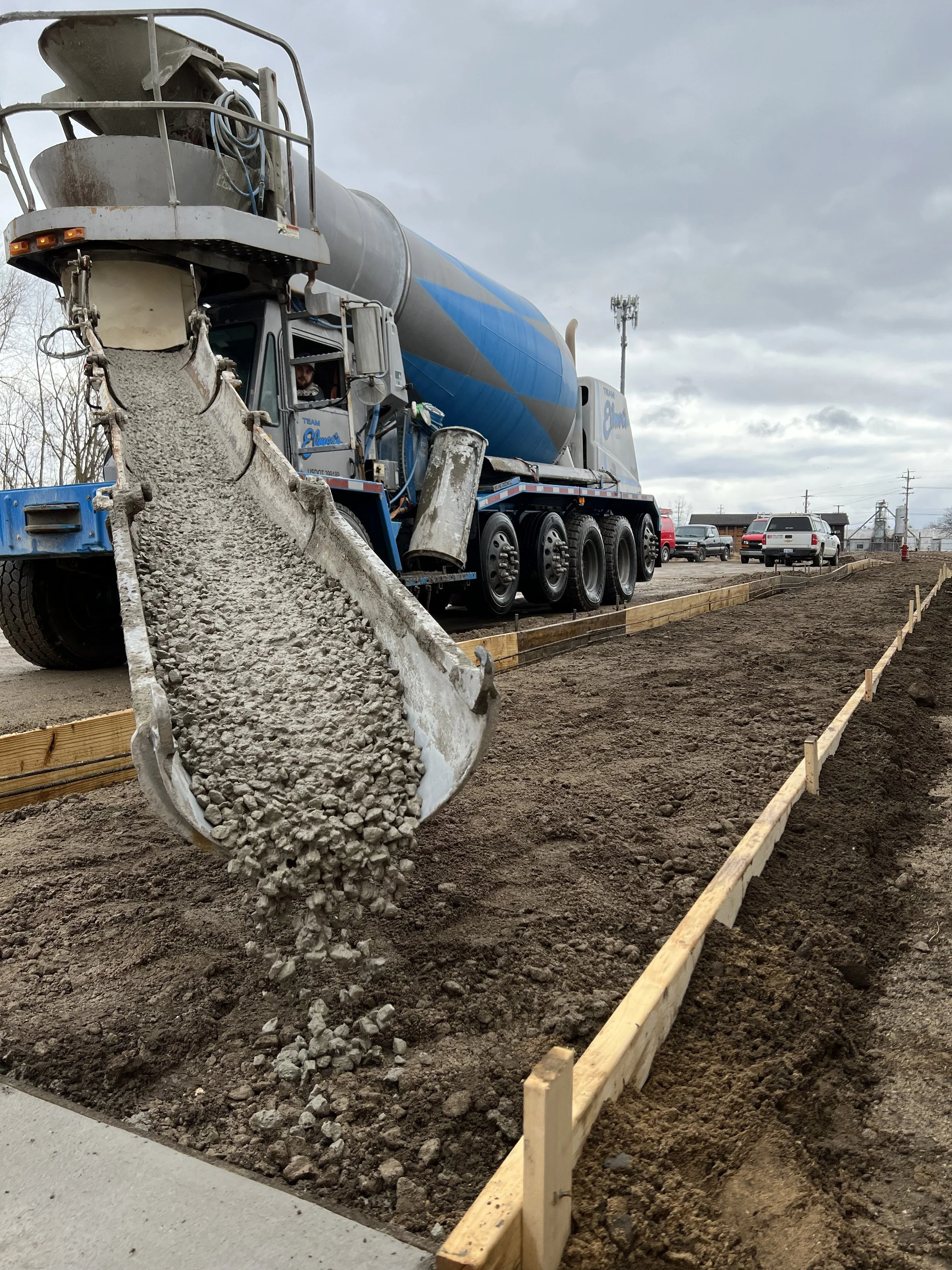 A concrete mixer truck pouring wet concrete into a designated area for construction, with wooden framing and a cloudy sky in the background.
