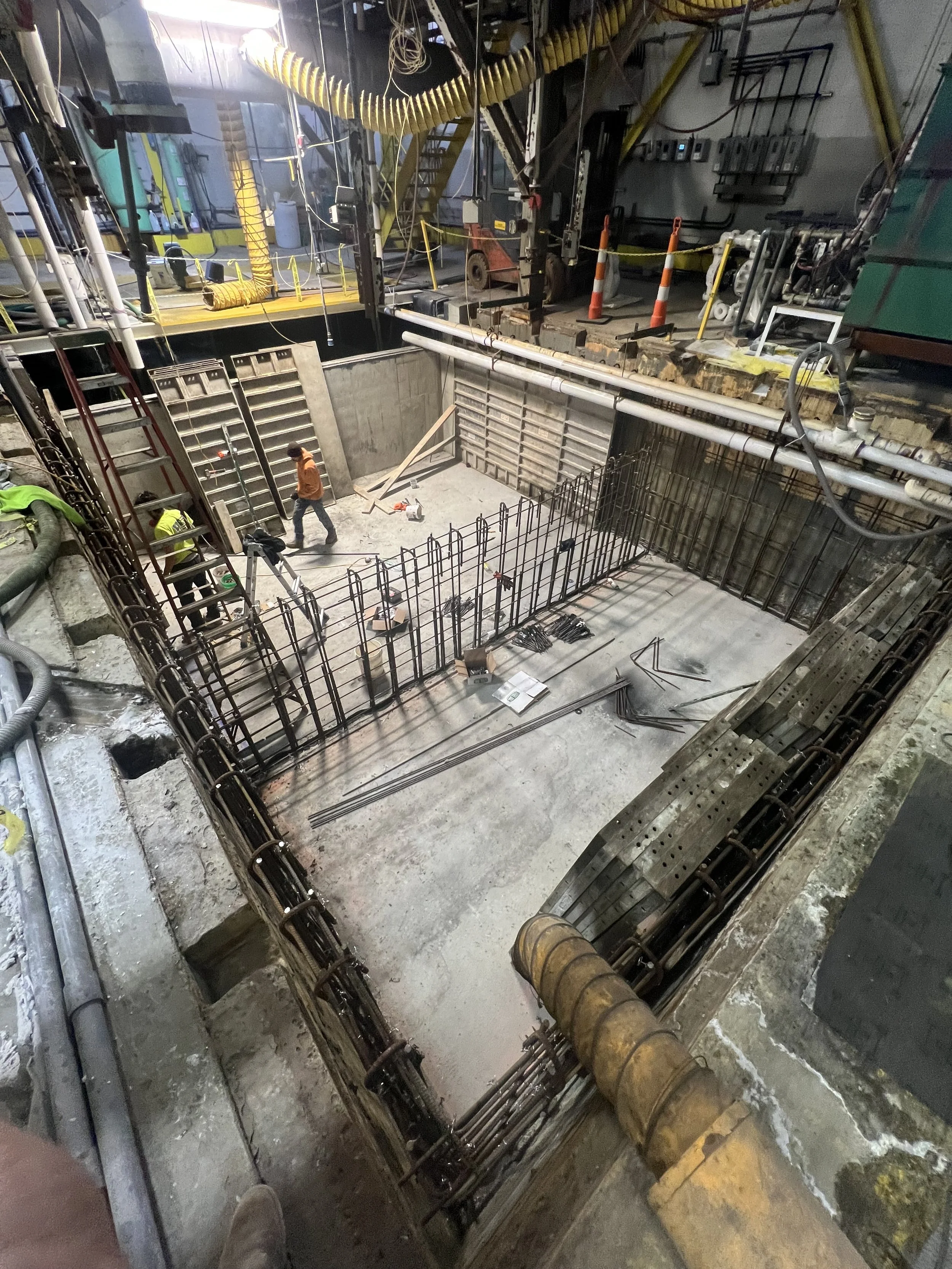 Construction workers working inside an elevator shaft, which is in the process of being built, with rebar reinforcing the walls and orange safety cones and barriers around the perimeter.