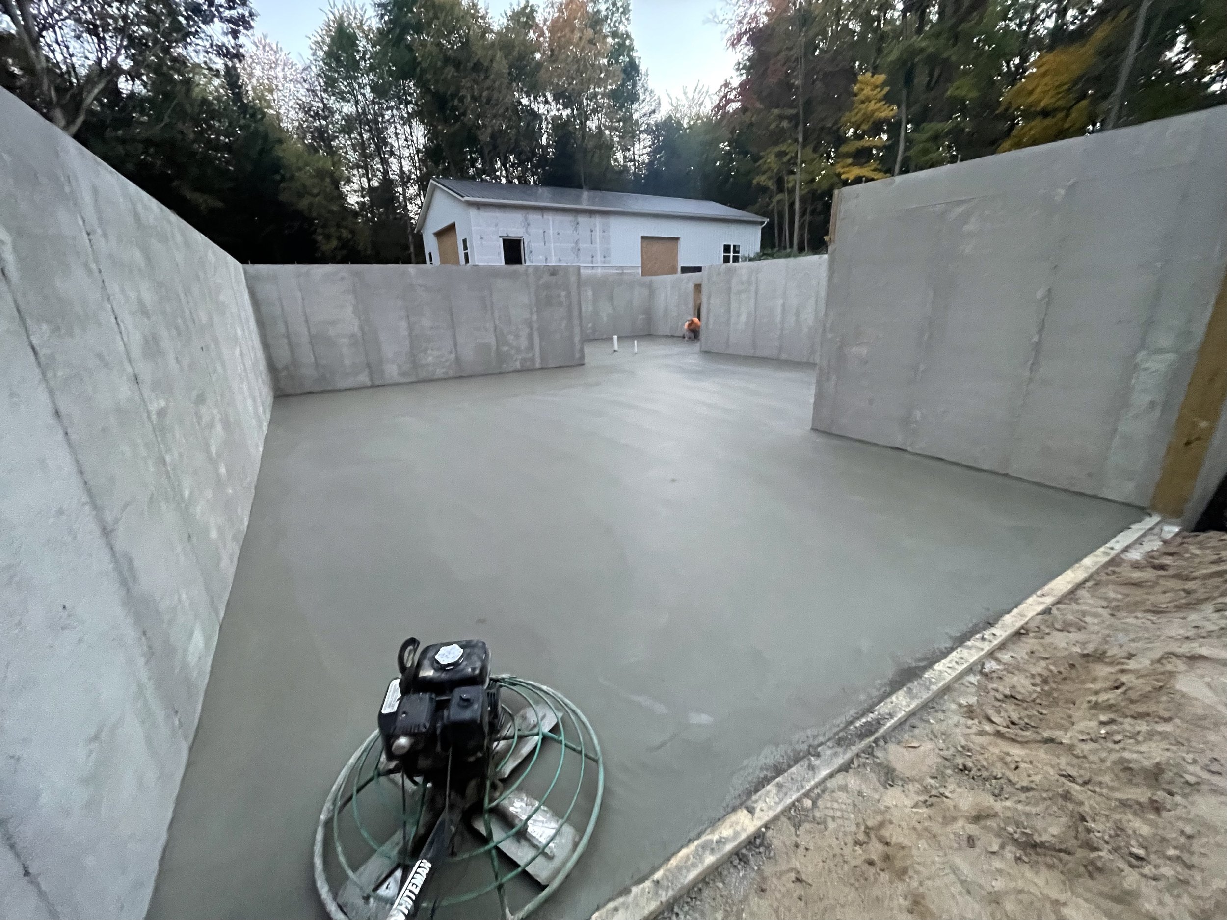 Concrete slab flooring in an outdoor construction site with concrete walls, a handheld power trowel, and a background of a partly built house and trees.