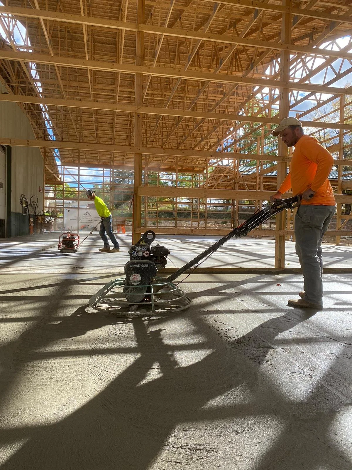 Two construction workers, one in an orange shirt and cap, and the other in a yellow shirt, laying down concrete inside a partially constructed wooden building.