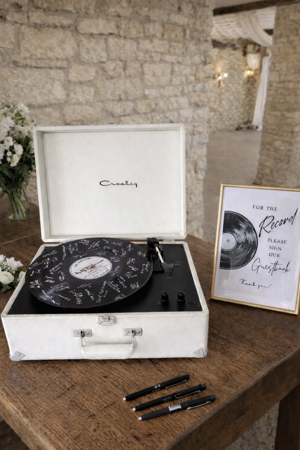 A vintage white record player with a black vinyl disc on it, surrounded by a sign requesting guests to sign the record at an event, set on a wooden table with pens nearby, inside a rustic stone-walled room.